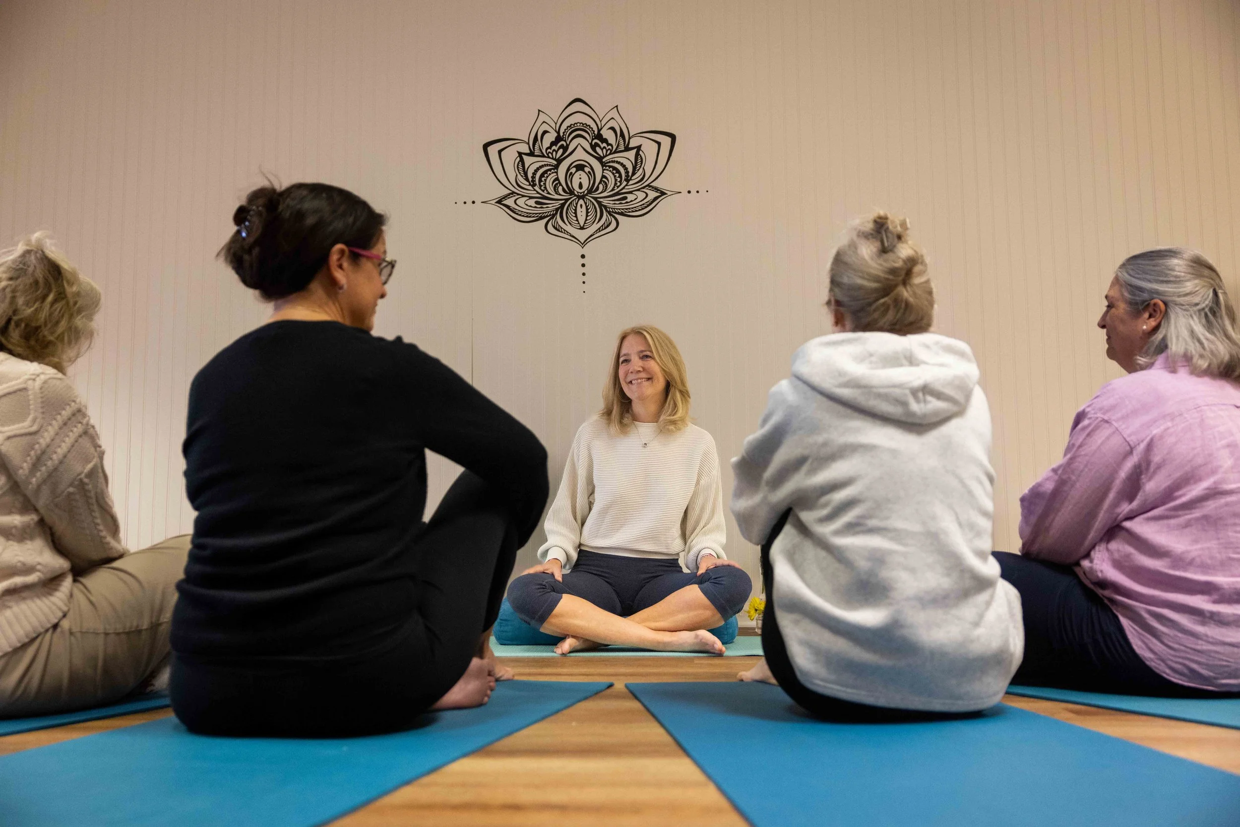 Group of women sitting in a circle on yoga mats, participating in a meditation session in a room with a lotus flower design on the wall.
