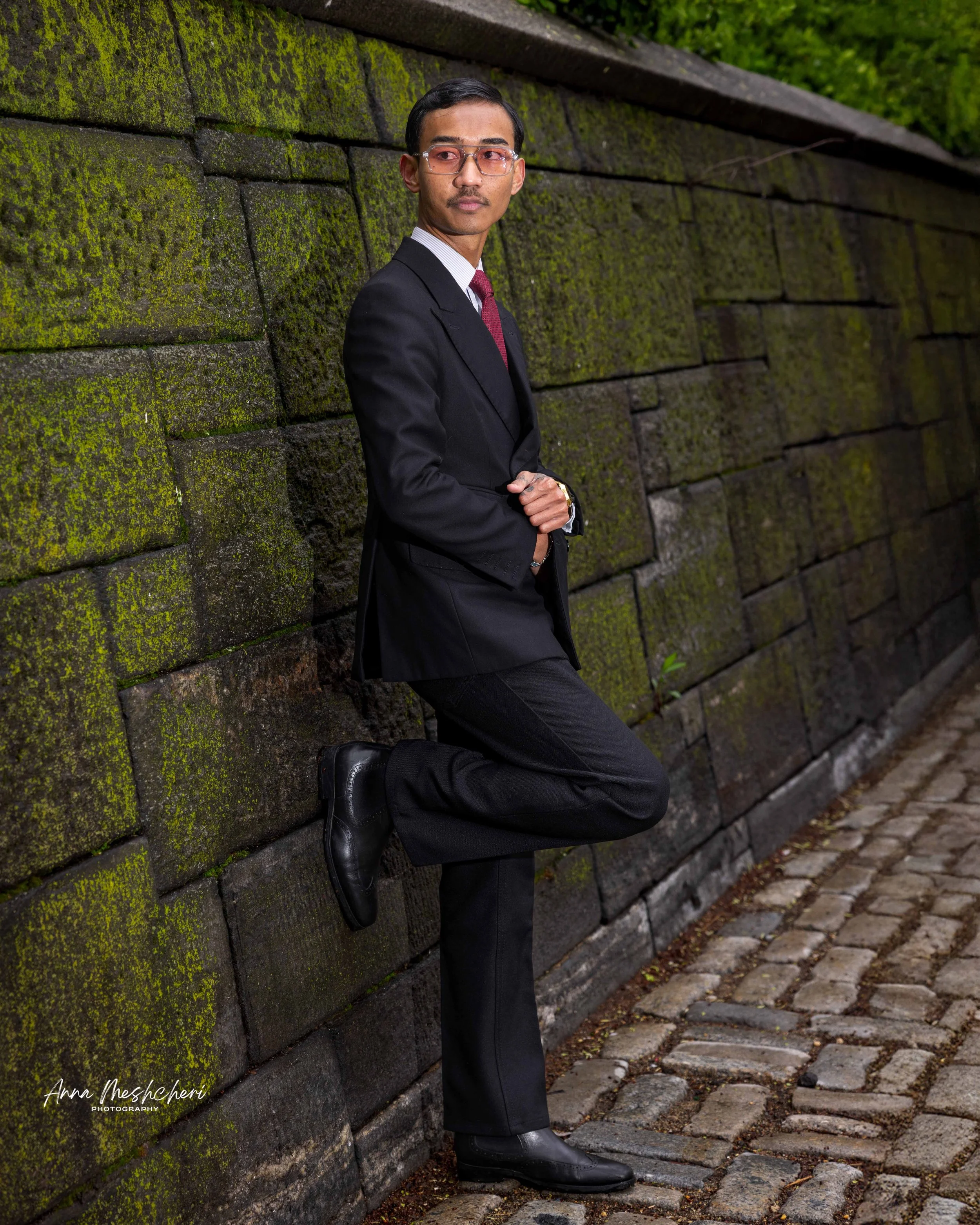 A man posing in a black suit near a wall covered with moss