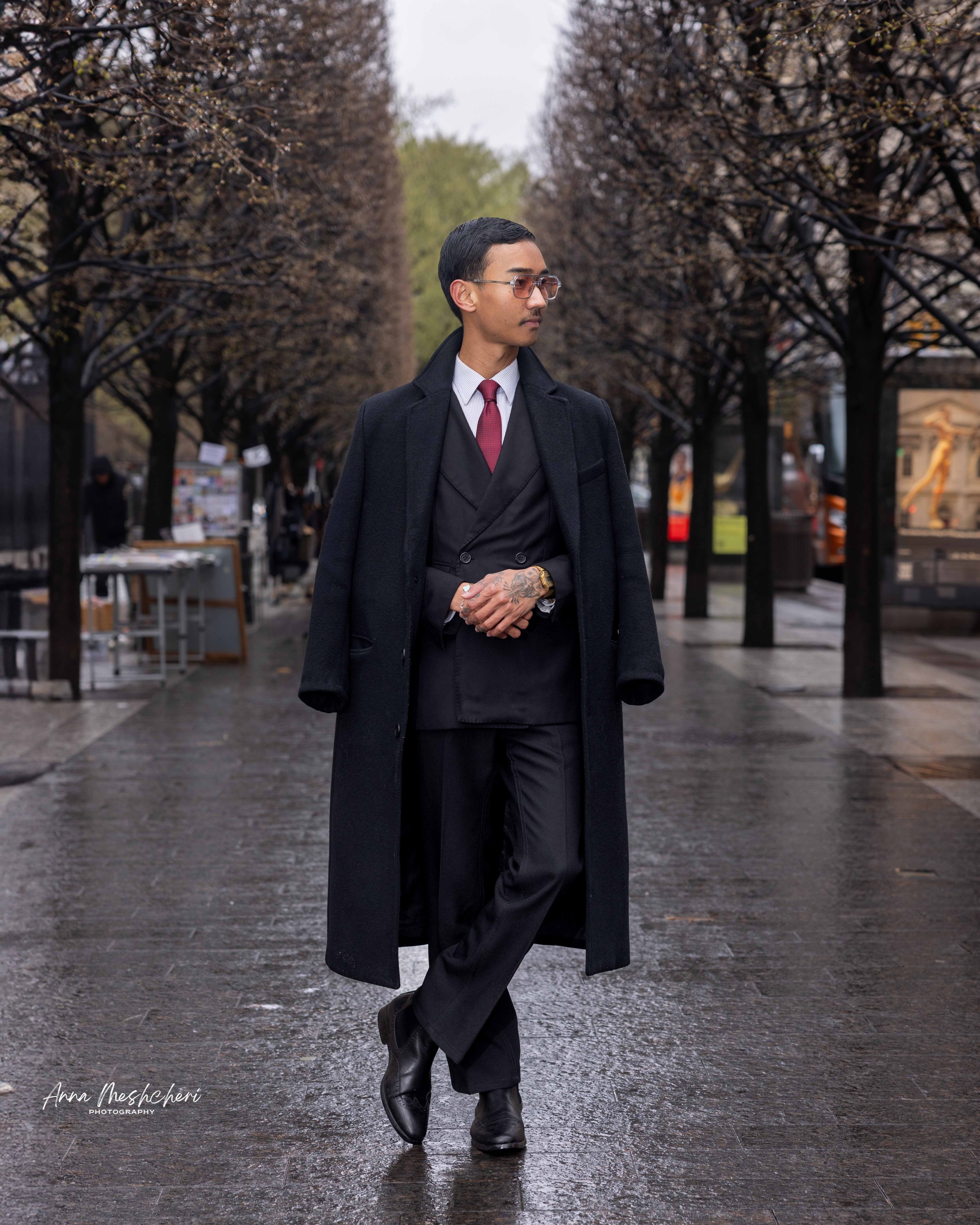 A man posing in a black suit and a black coat in an alley near the Metropolitan museum of art