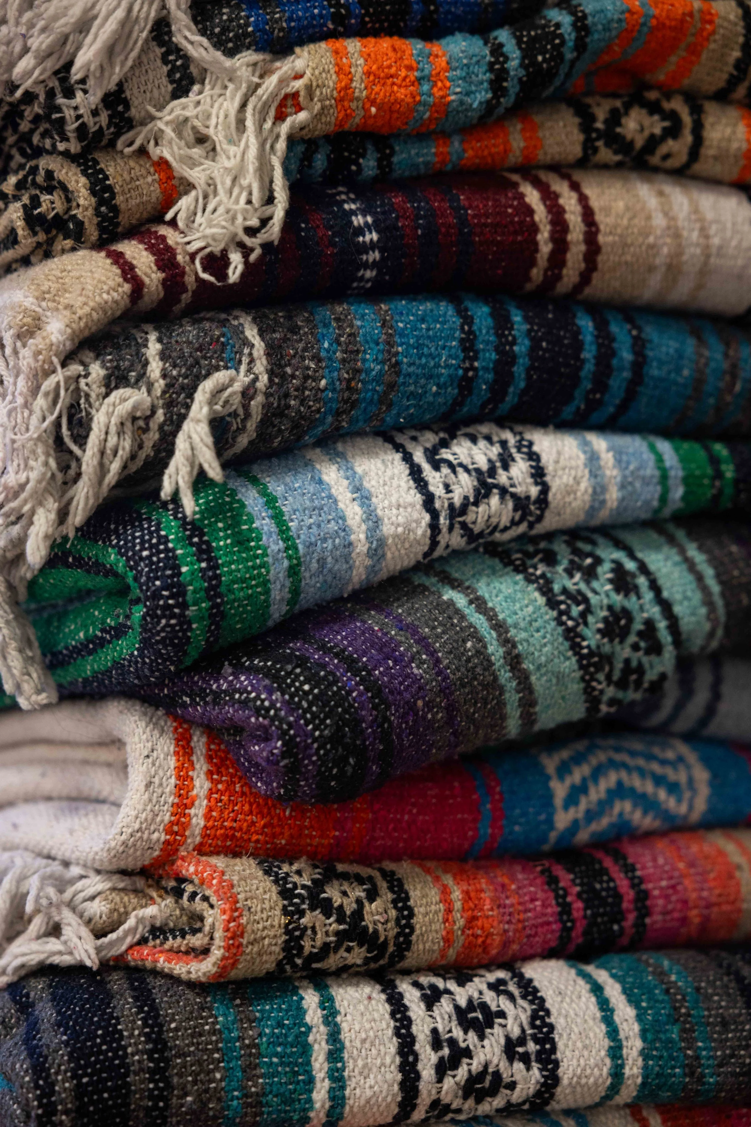 Stack of folded textiles or blankets with colorful, intricate patterns and fringed edges, arranged in horizontal layers.