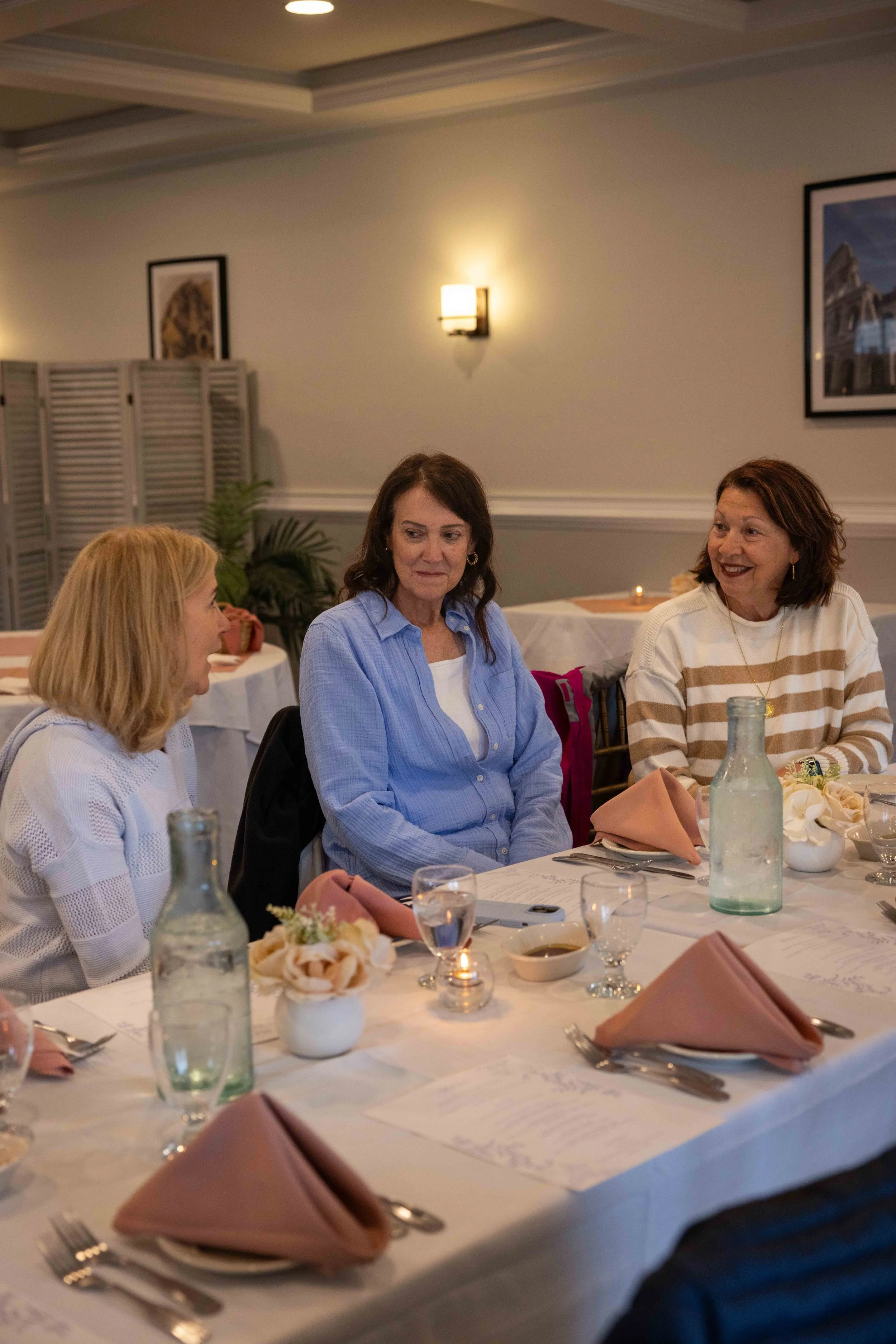 Women sitting at a round dining table with pink napkins, floral centerpieces, glass water bottles, and cutlery in a well-lit room, engaged in conversation.