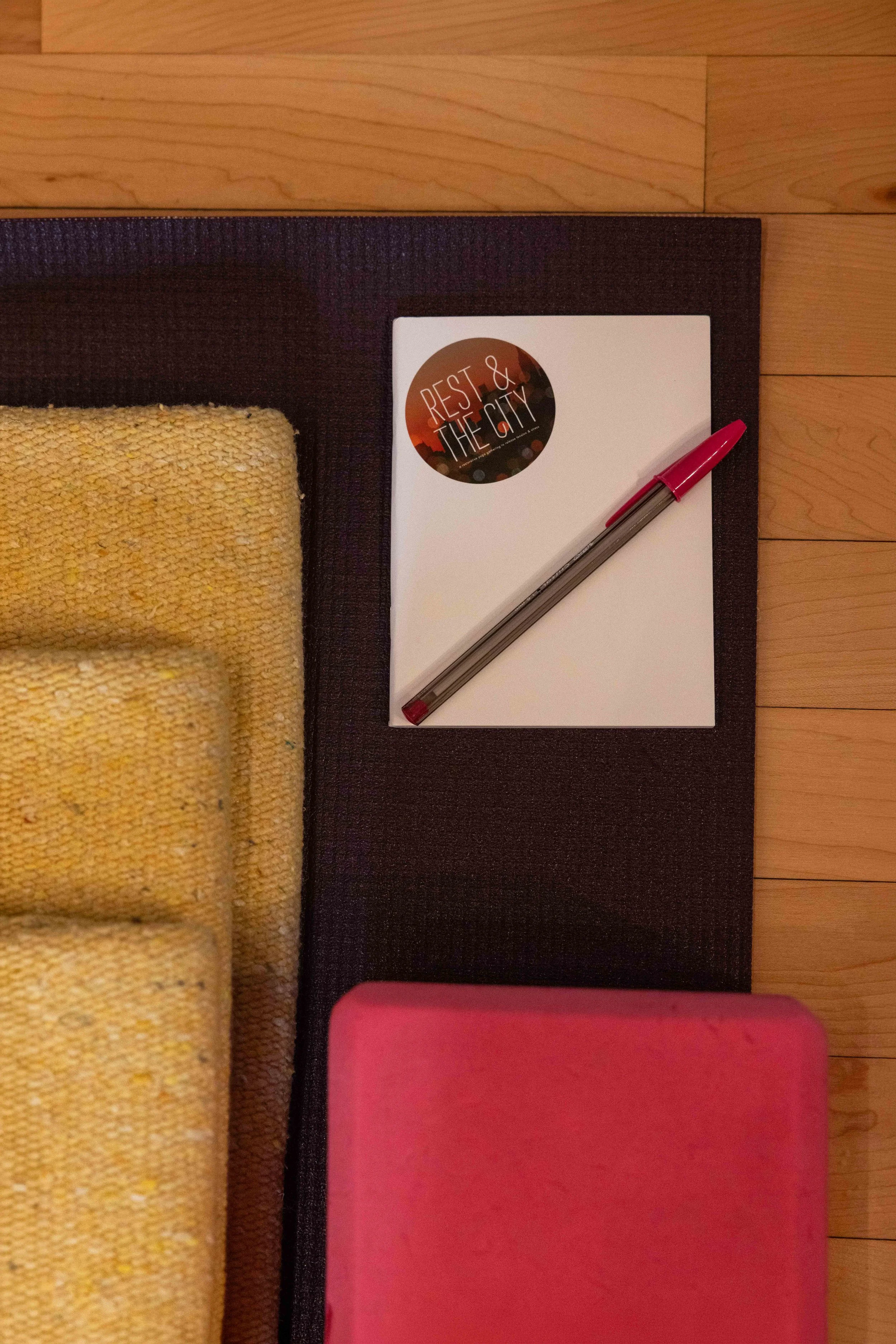 A table with a yellow towel, a dark mat, a white notebook with a sticker that says 'Rest & The City,' a pink pen, and a pink item on a wooden floor.