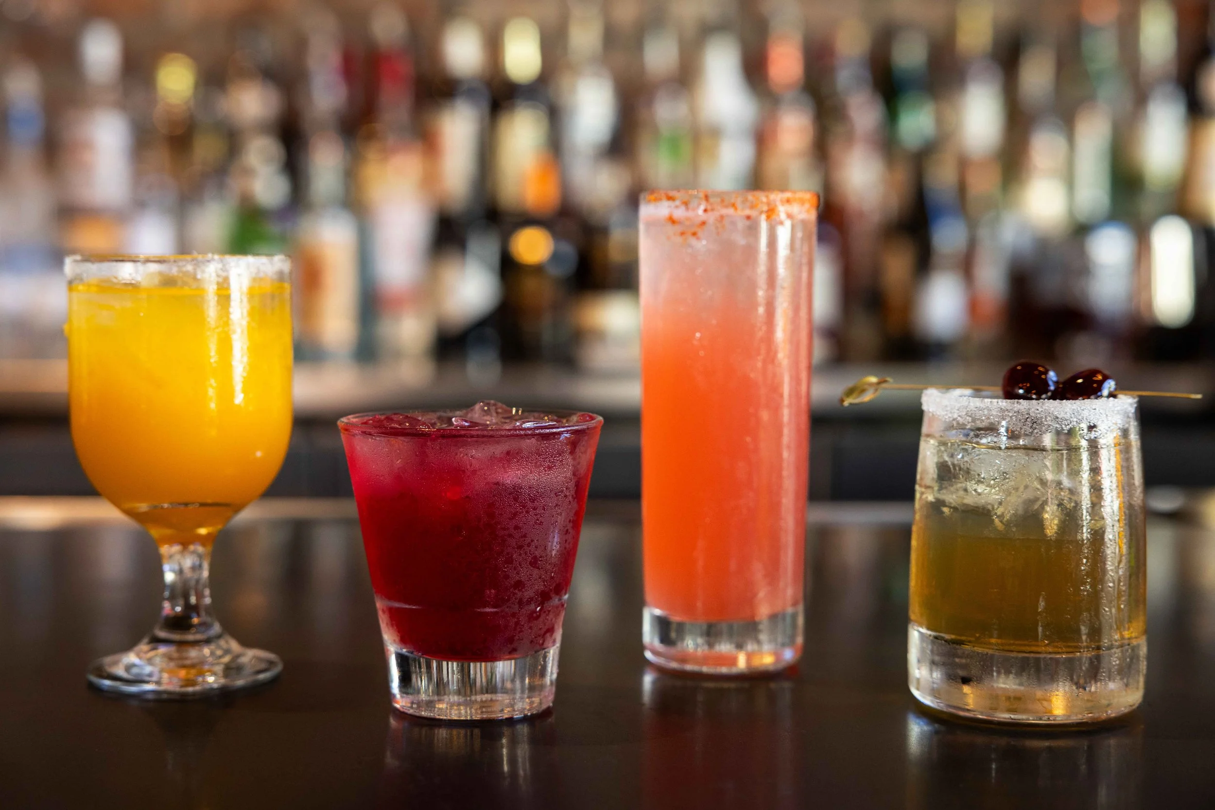 Four colorful cocktails on a bar counter with a blurred bar background.