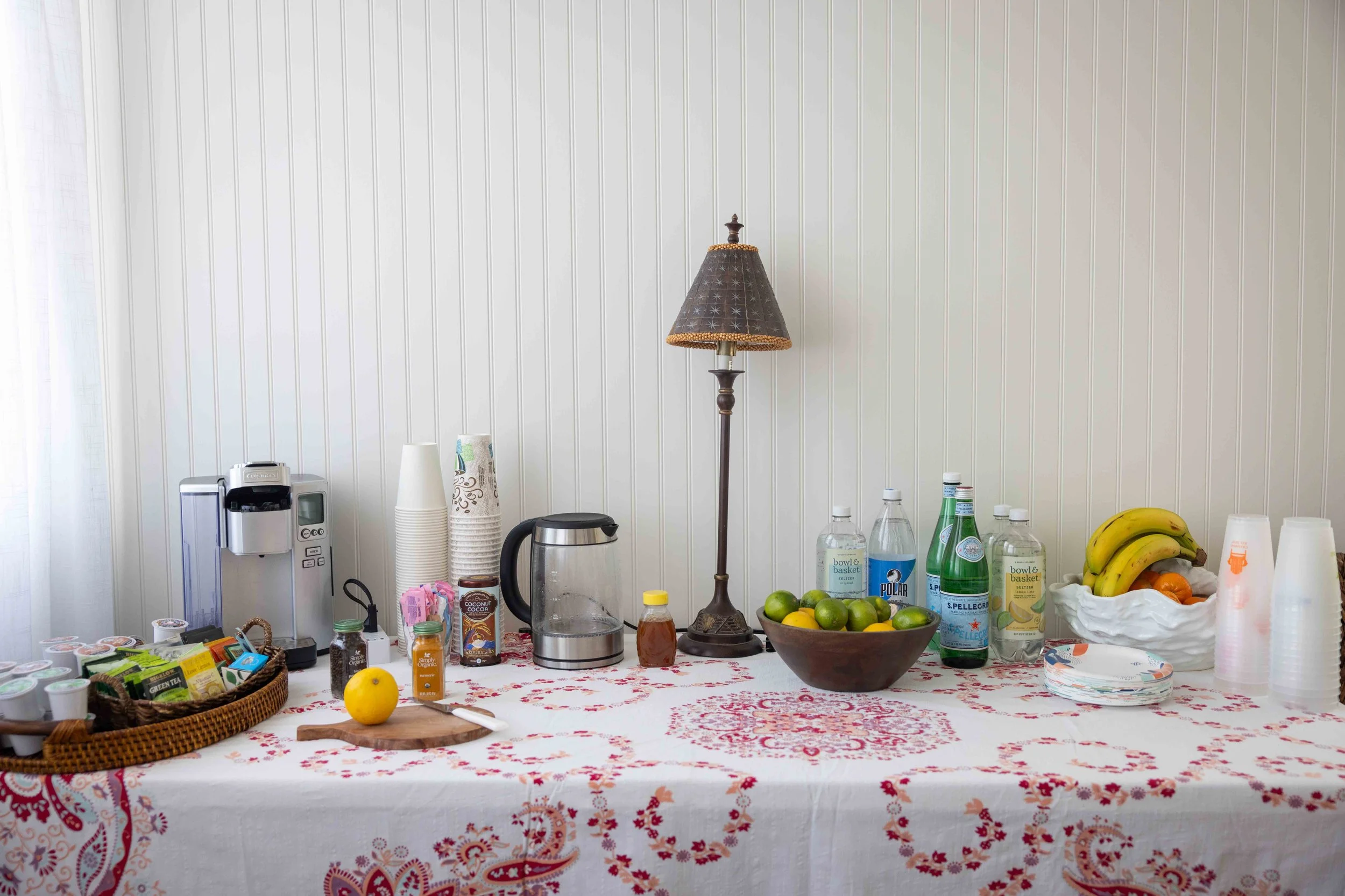 Table with coffee, drinks, fruit, and snacks in front of a white wall with vertical paneling.
