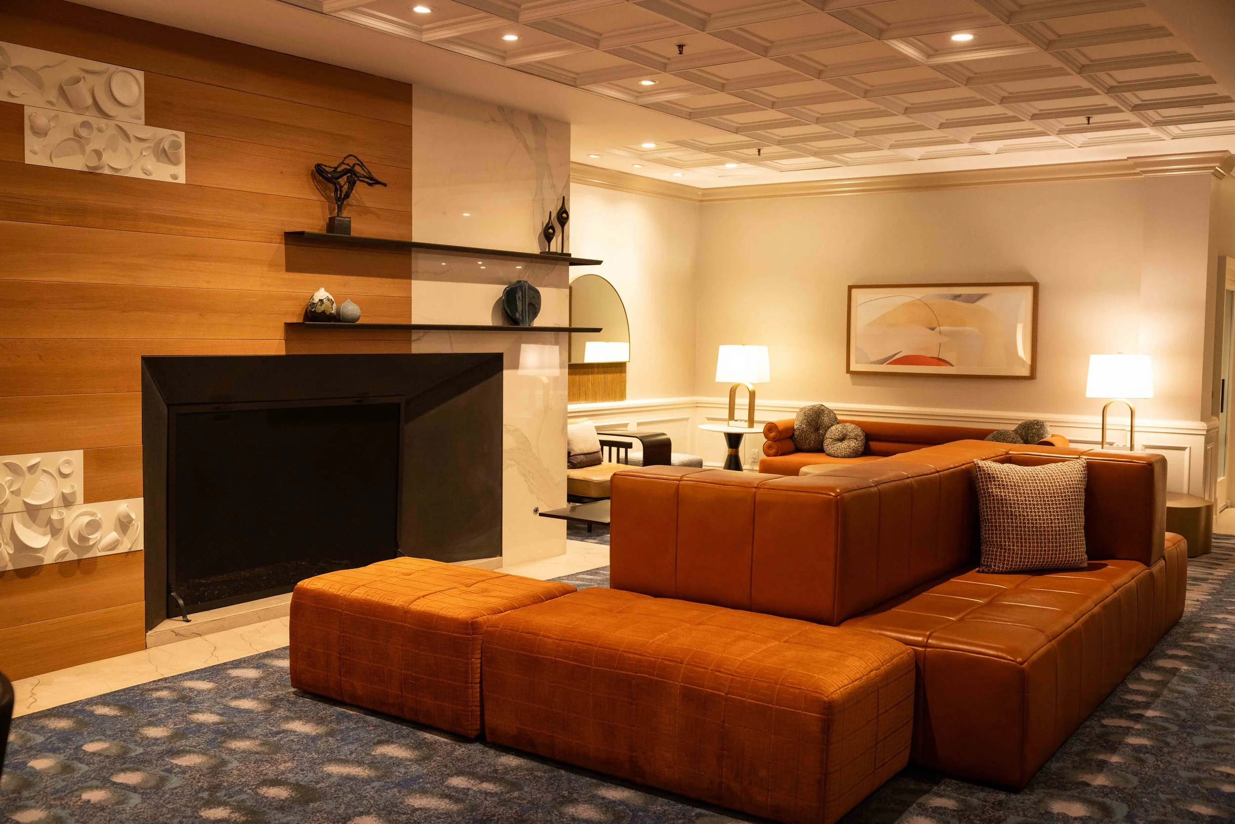Living room with a brown leather sofa, beige and grey patterned pillows, two lamps, abstract artwork, and shelves with decorative objects on a wooden and marble wall.