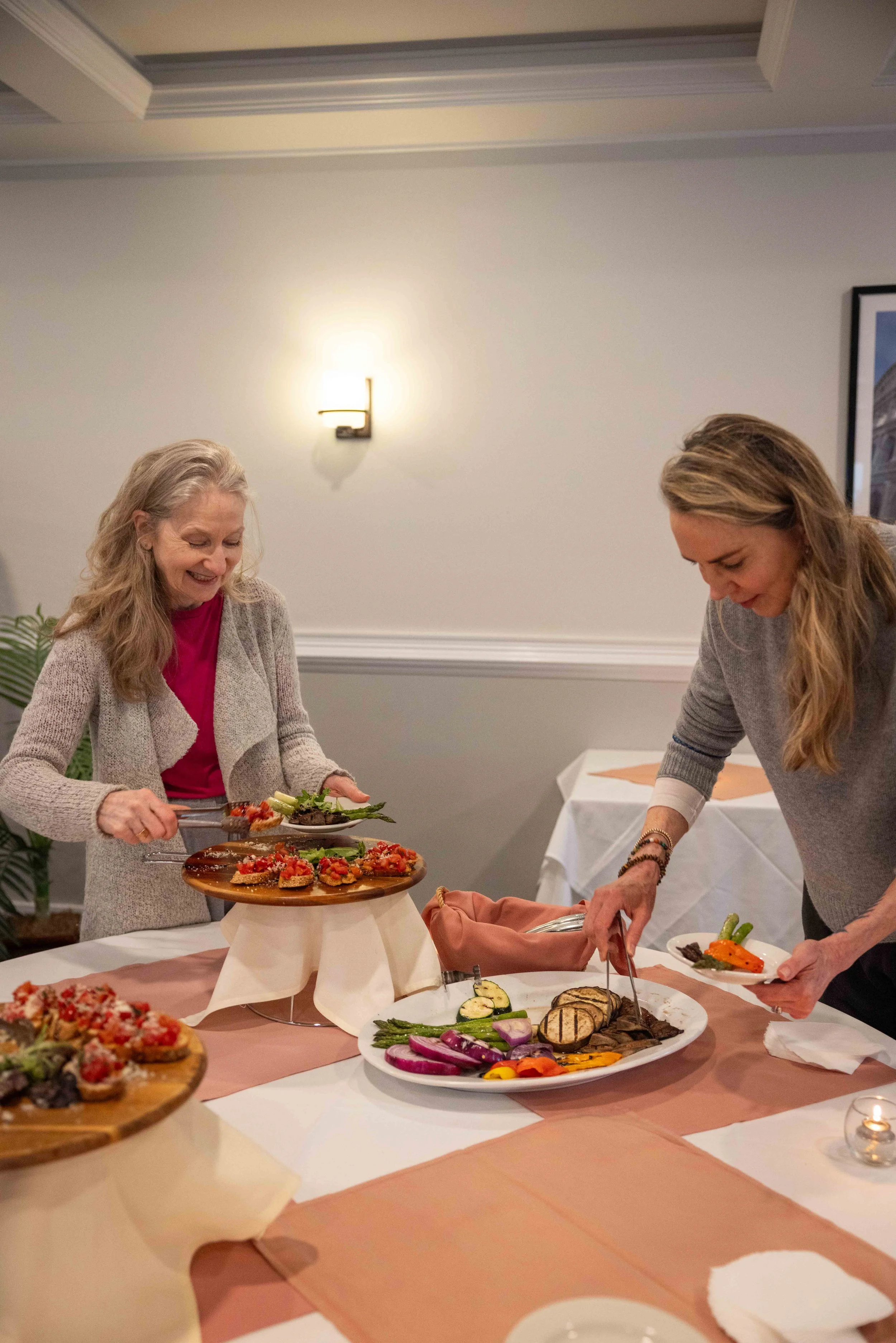 Two women are serving themselves food from a buffet table decorated with a pink tablecloth and a beige runner. The table has various dishes including grilled vegetables and appetizers, with a wooden serving tray on a stand. One woman has long blonde 