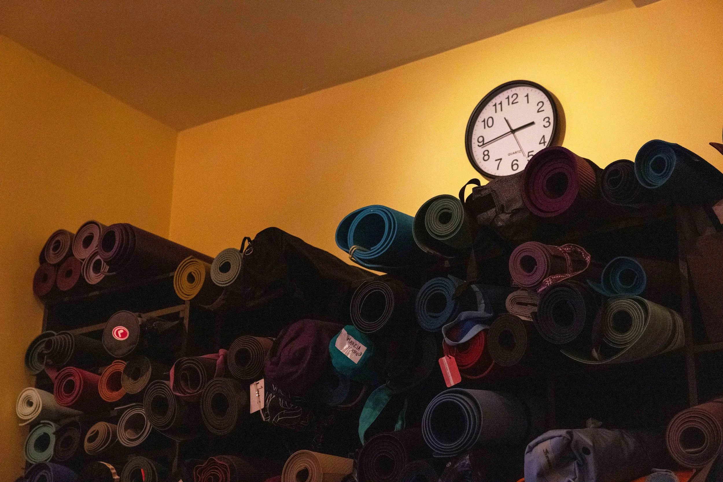 Stacked yoga mats in various colors on shelves against a yellow wall with a round wall clock showing approximately 2:15.