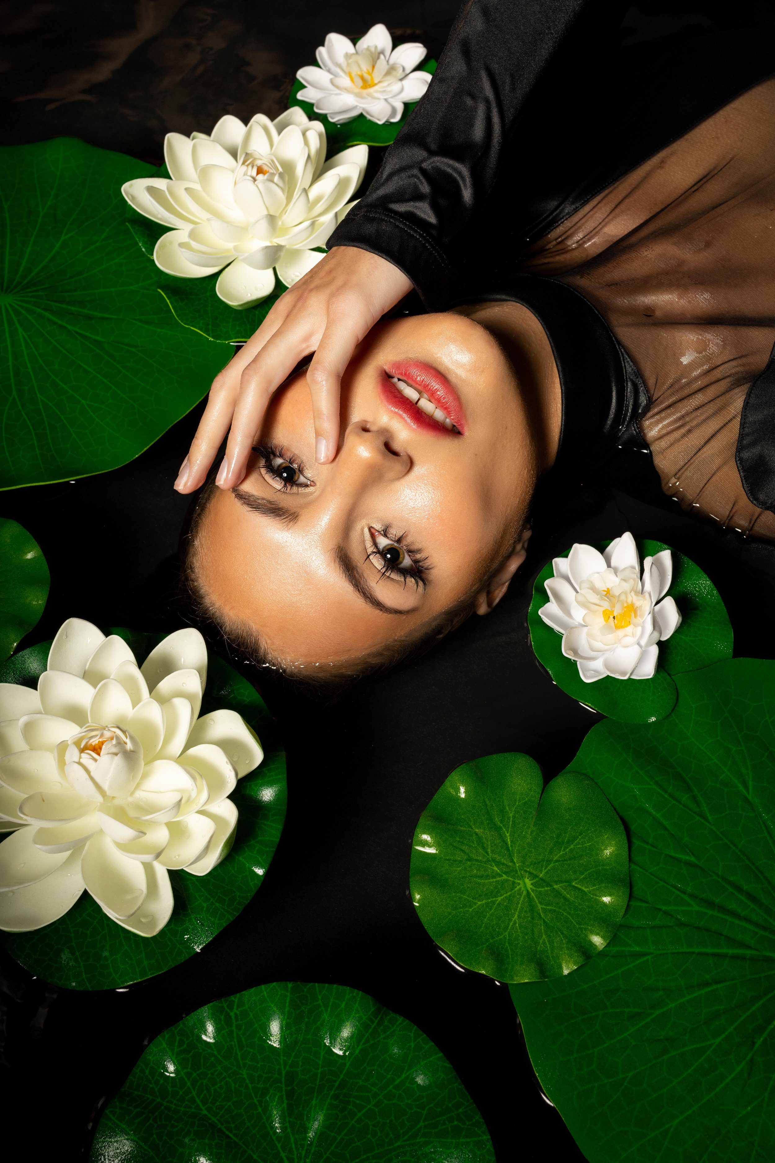 Woman lying in water surrounded by white water lilies and green lily pads, with one hand on her face.