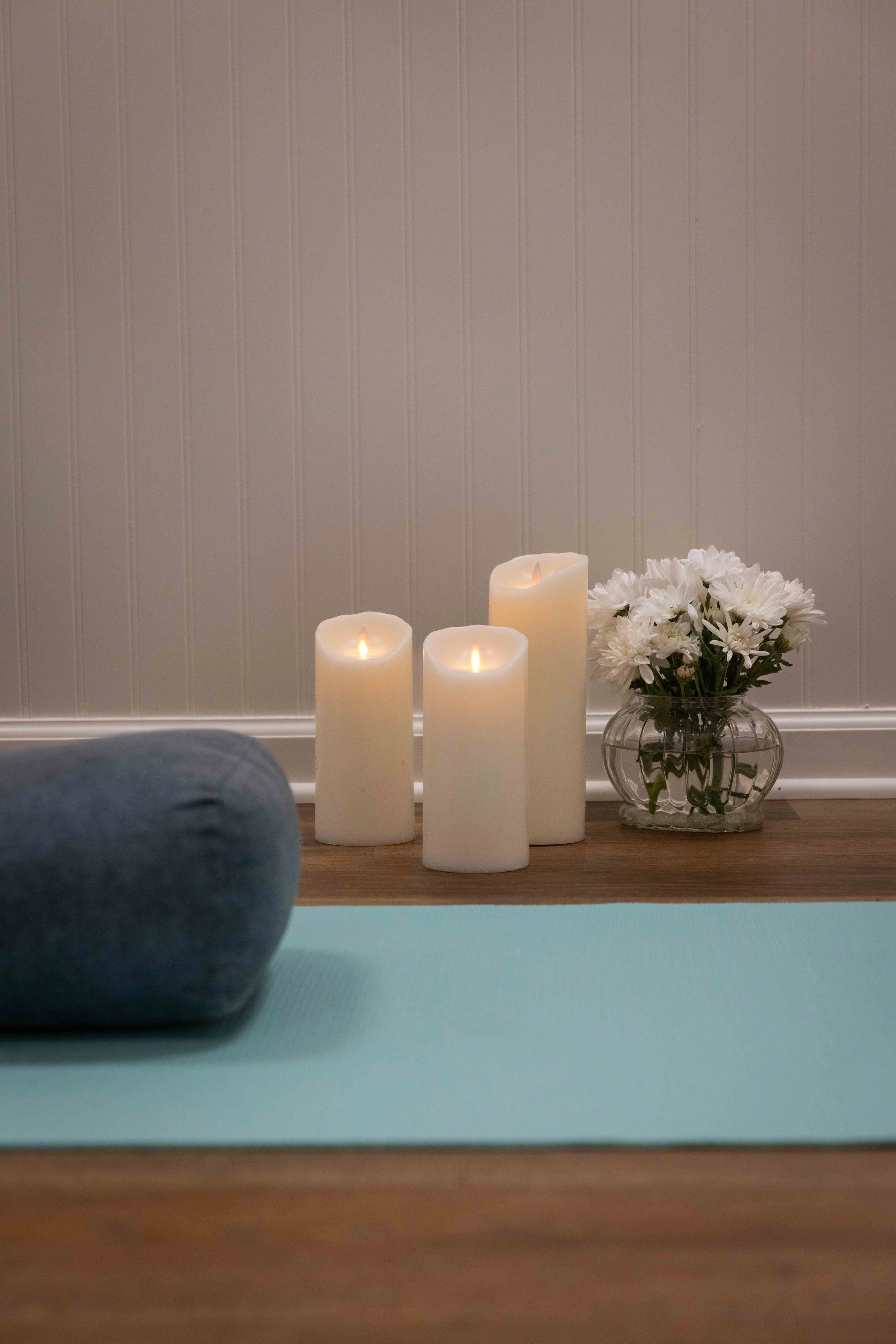 View from a yoga mat showing a person's leg, with candles, white flowers in a glass vase, a white wall with vertical lines, and a hardwood floor.
