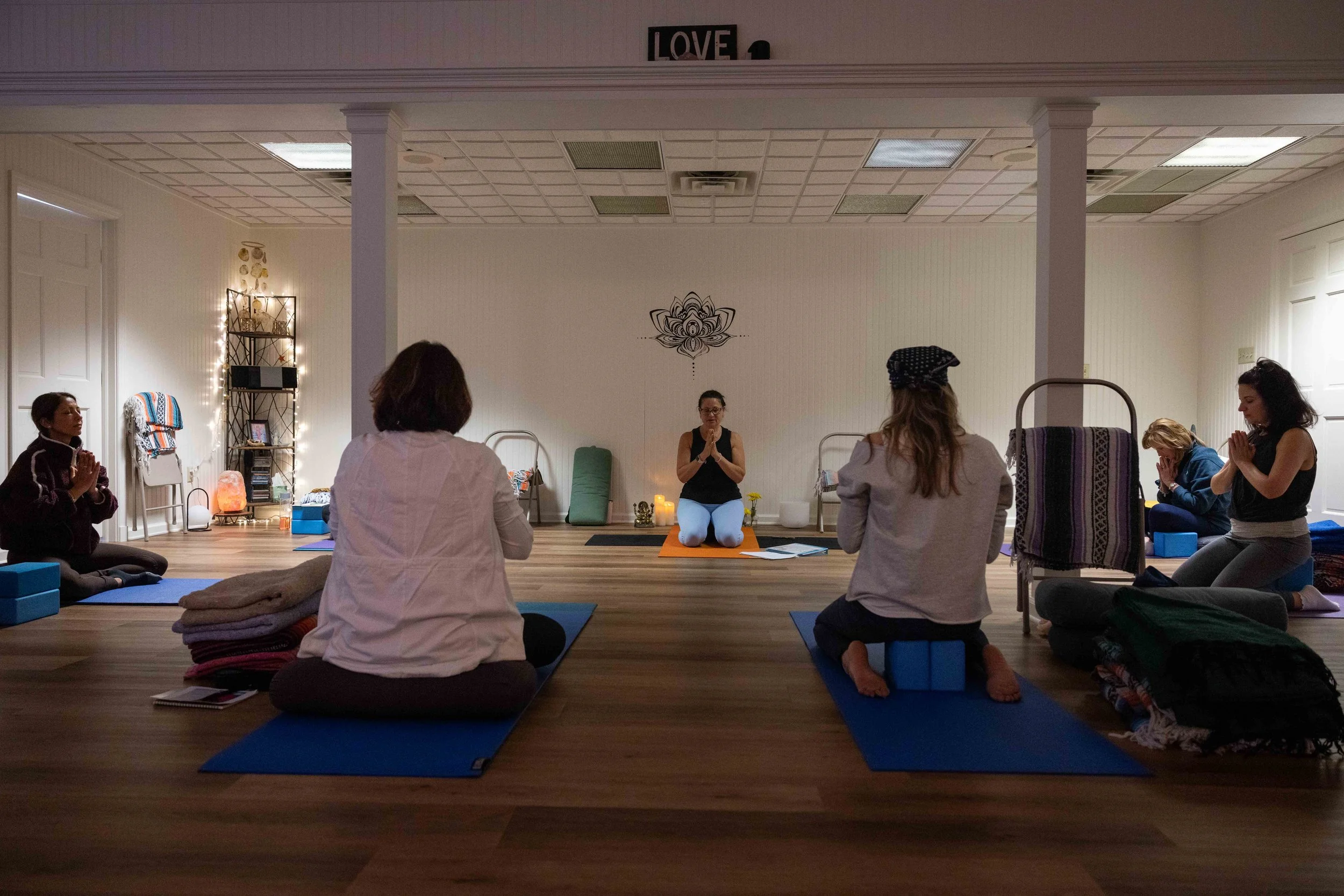 A group of people participating in a yoga or meditation class in a softly lit, cozy indoor space. The instructor is seated on a yoga mat at the front, leading the session, while participants sit cross-legged or kneel on mats with closed eyes and hand