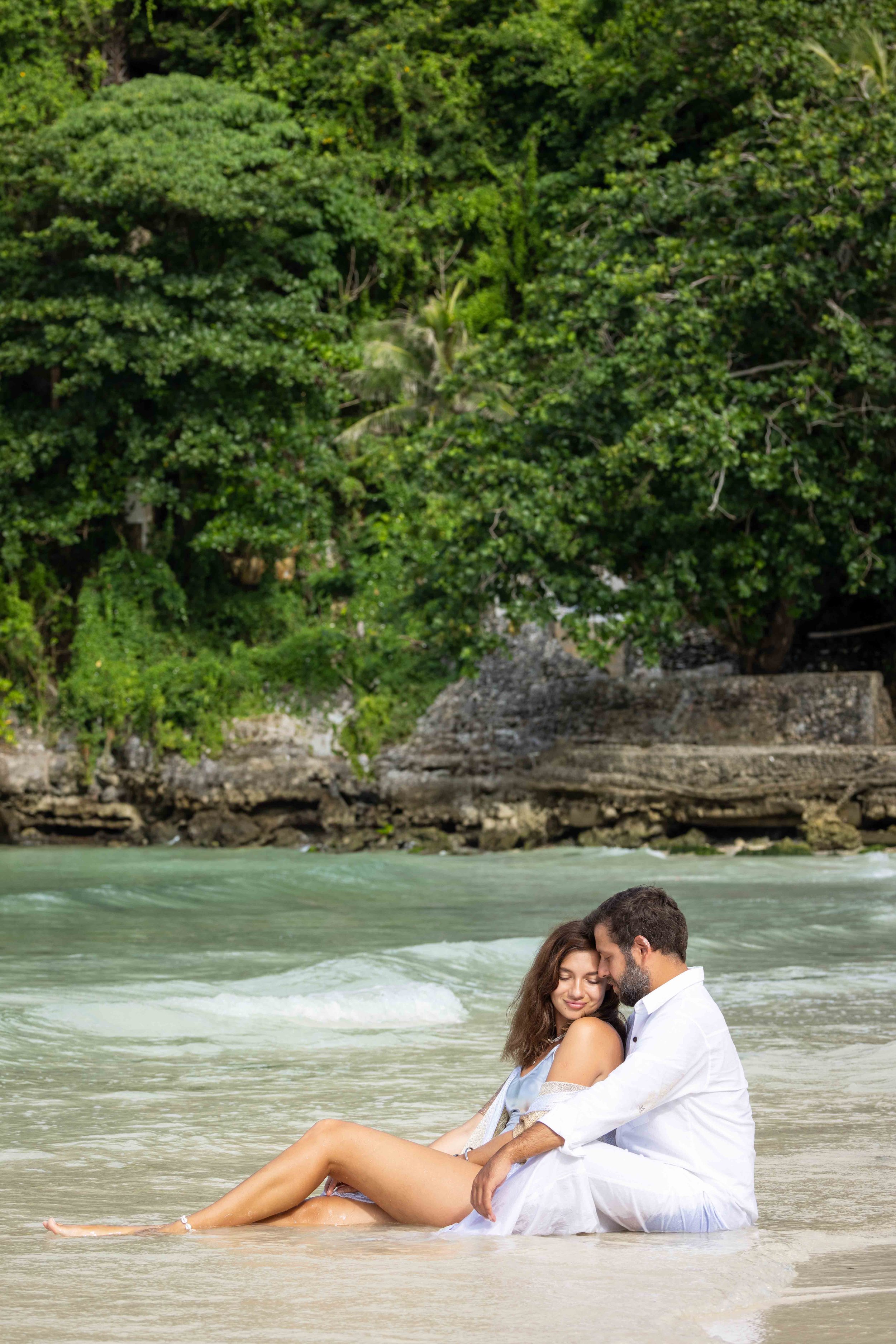 Couple sitting on a beach with clear water and lush green trees in the background. Boracay beach lifestyle family photoshoot by Anna Meshcheri