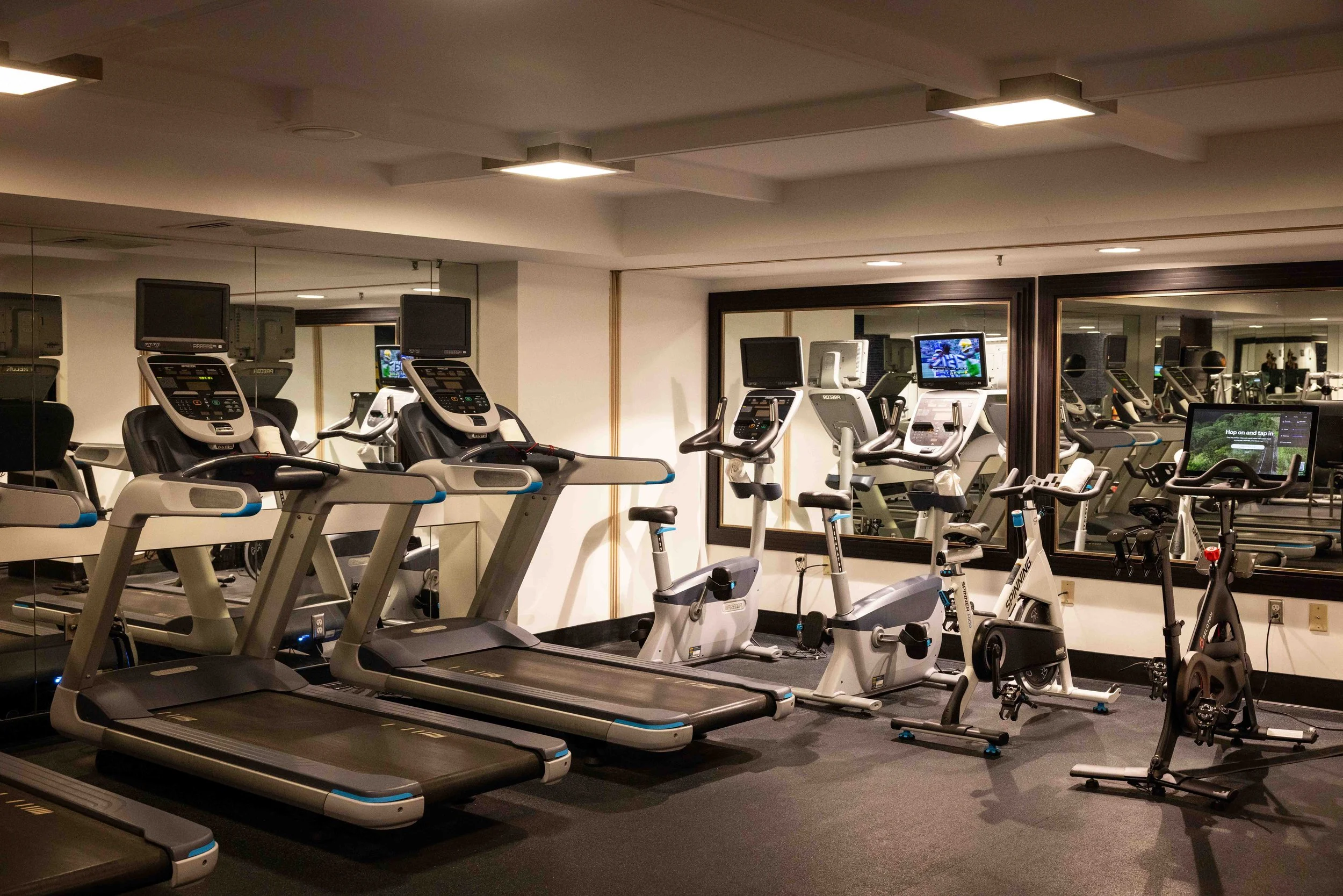 A fitness room with several treadmills, stationary bikes, and elliptical machines, with a large mirror on the wall reflecting the workout equipment and space.
