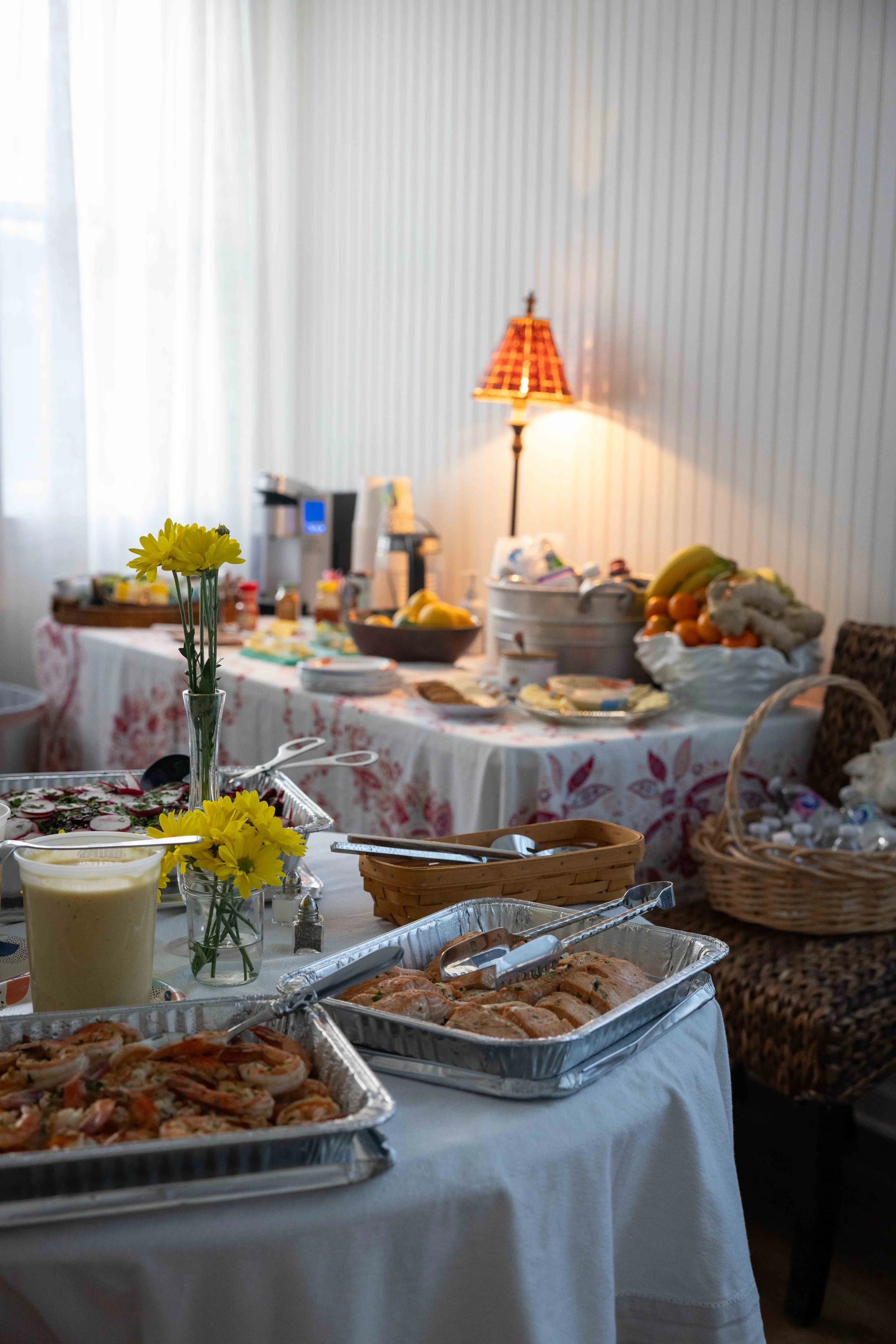 A table set for a gathering with trays of snacks including pizza, a bowl of fruit, and a yellow beverage, with a vase of yellow flowers in the foreground. In the background, there is a lamp and a buffet table with various foods and kitchen appliances