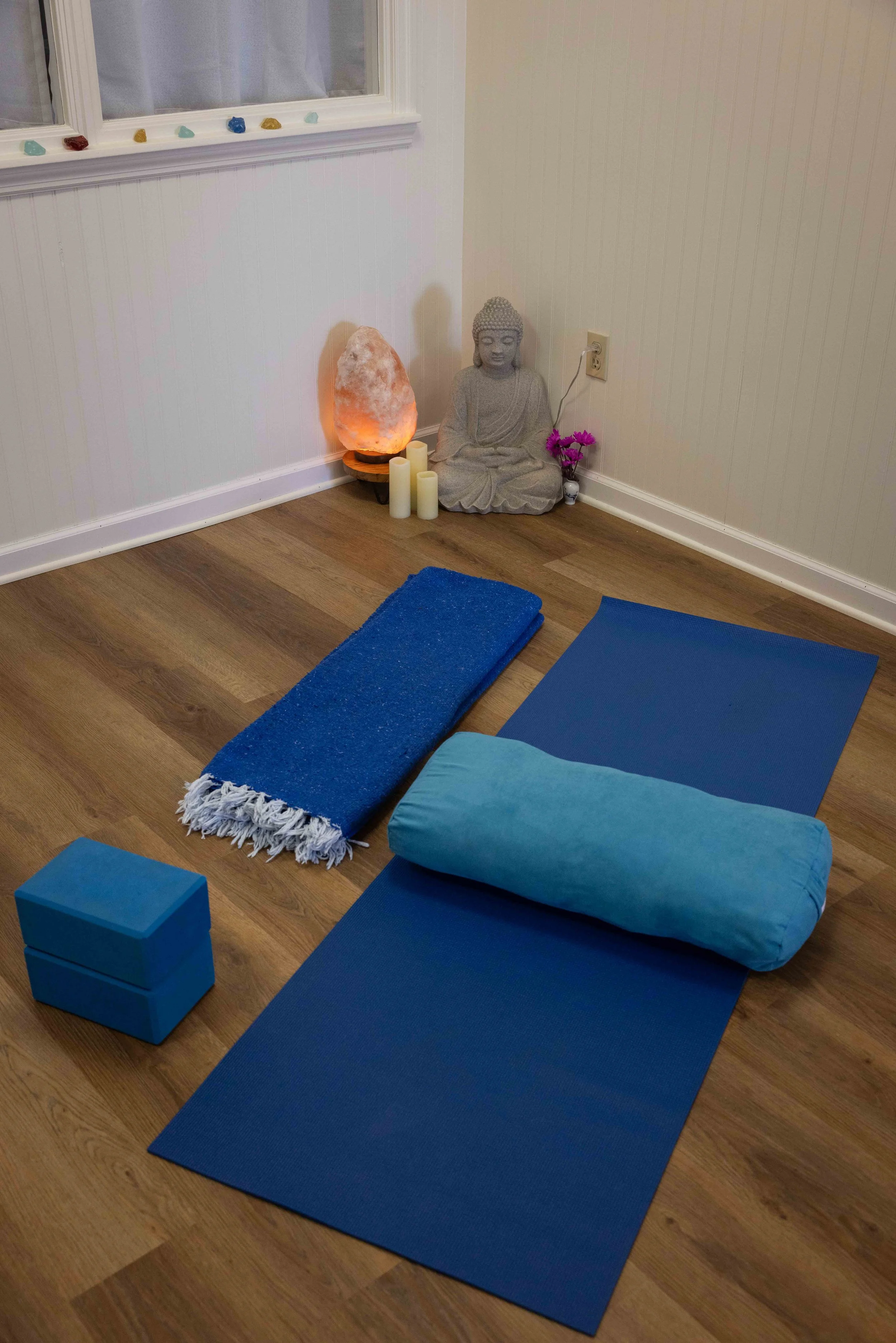 Yoga mats, a bolster cushion, a salt lamp, candles, a Buddha statue, and flowers in a corner of a room with wooden floor and a window.