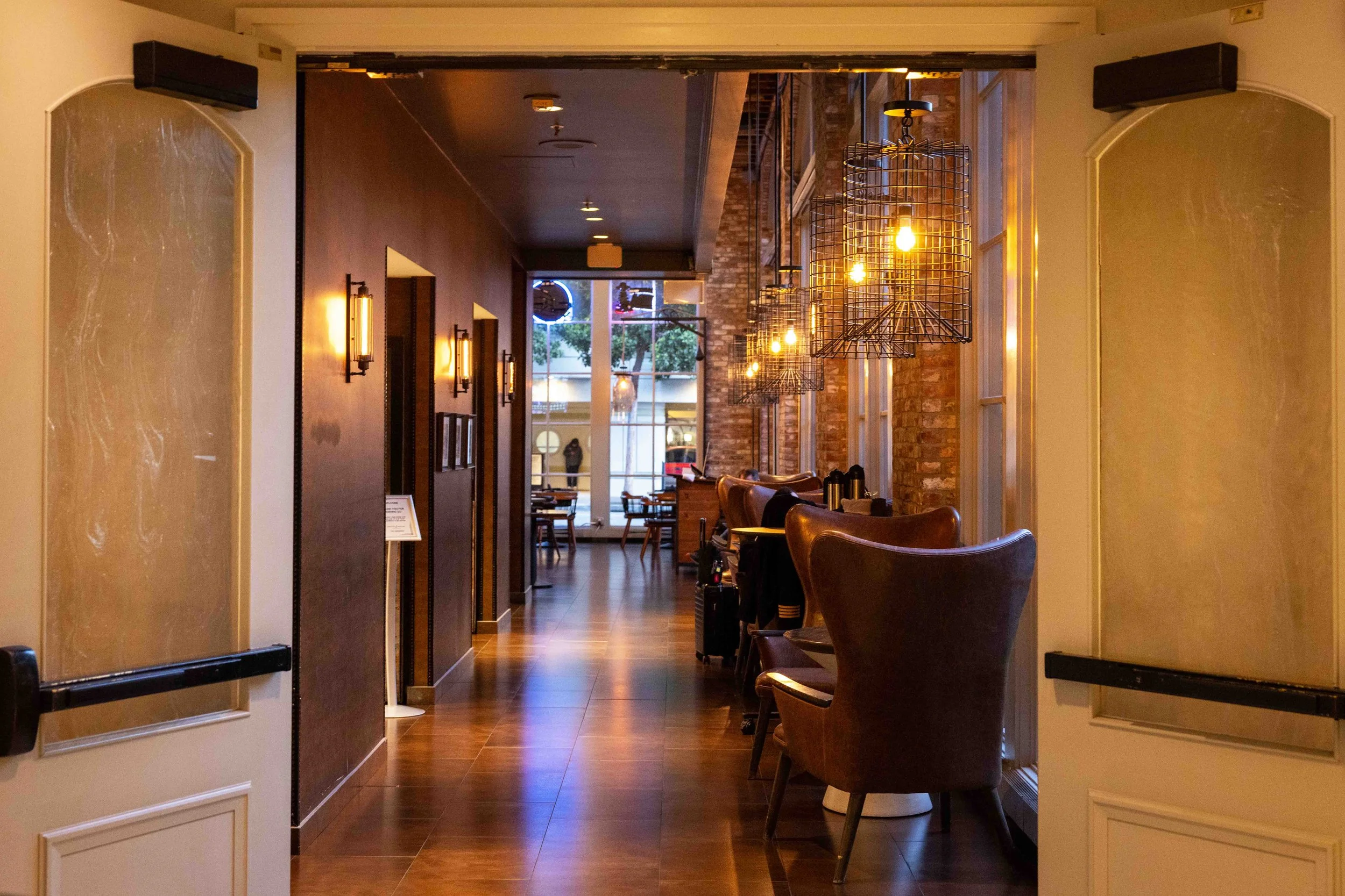 Interior of a cozy restaurant or cafe with warm lighting, wooden floors, leather chairs, brick wall accents, and large windows showing trees outside.