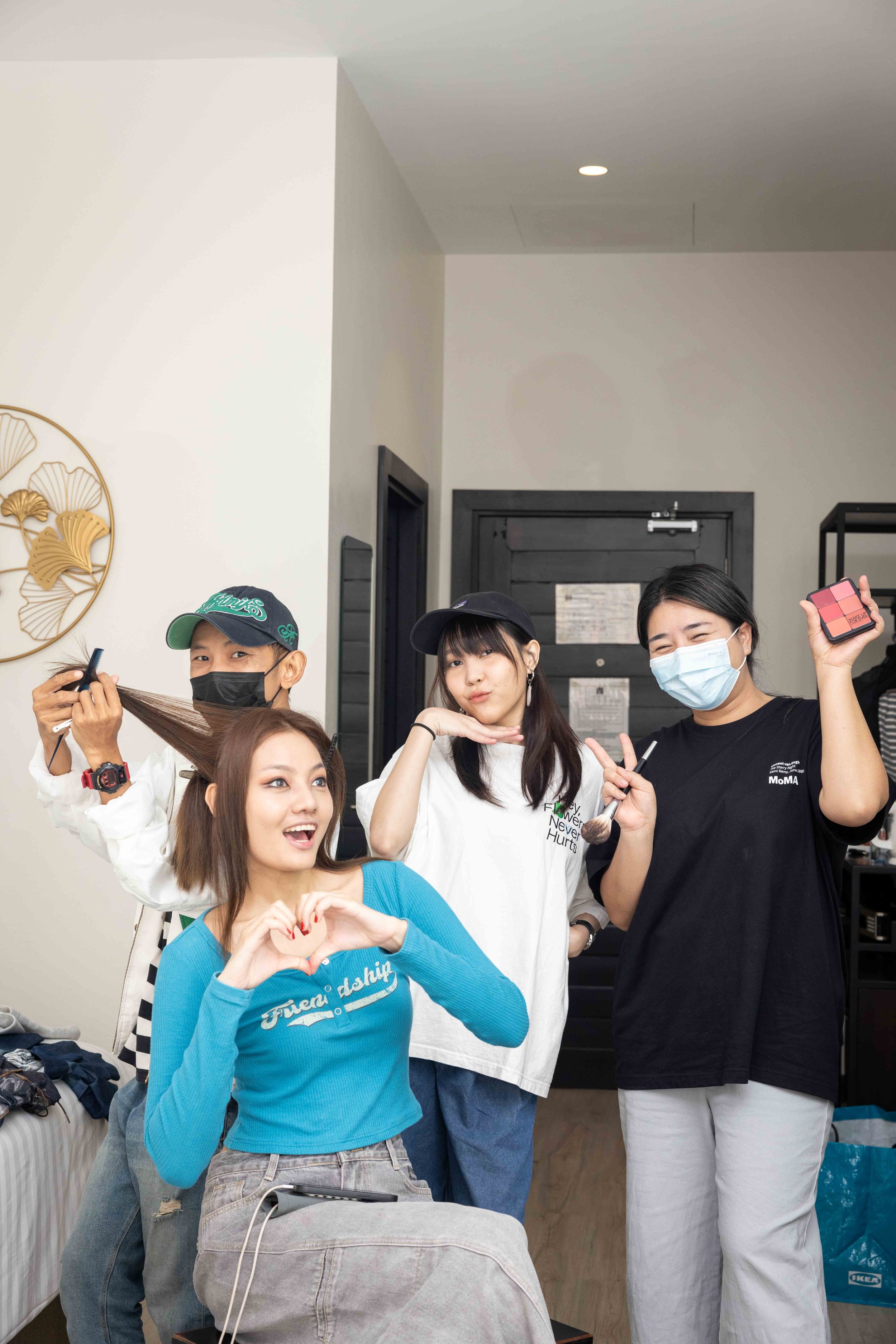 A group of four people in a room, with one sitting in a chair. The person seated in the center, wearing a blue top, is being attended to by three others, one holding a comb and working on their hair, while the others stand nearby smiling or making ge