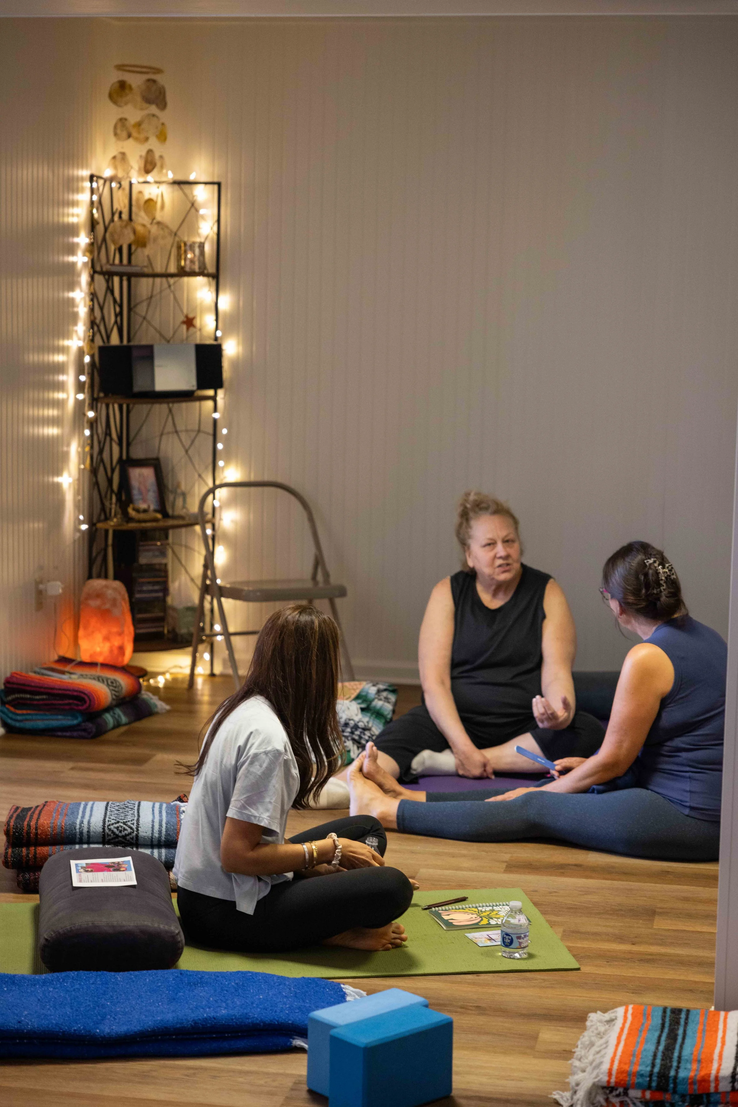 A small group of women participating in a yoga or meditation class in a cozy room decorated with string lights, framed pictures, and a salt lamp. They are sitting on yoga mats and cushions, engaging in conversations and practices.