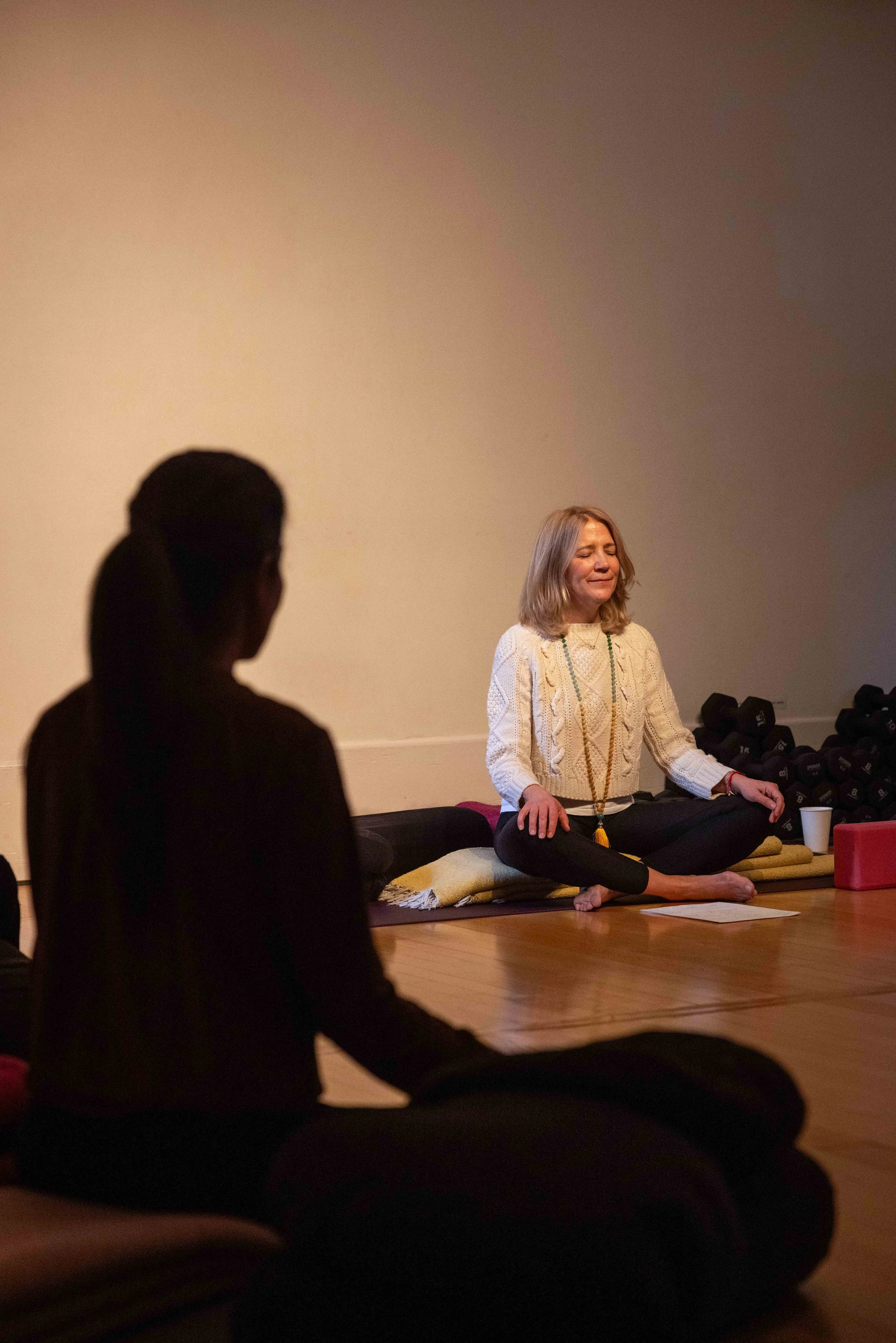A woman with shoulder-length blonde hair sitting cross-legged on a meditation cushion with her eyes closed, practicing meditation in a dimly lit room, with some dumbbells and a yoga block nearby.