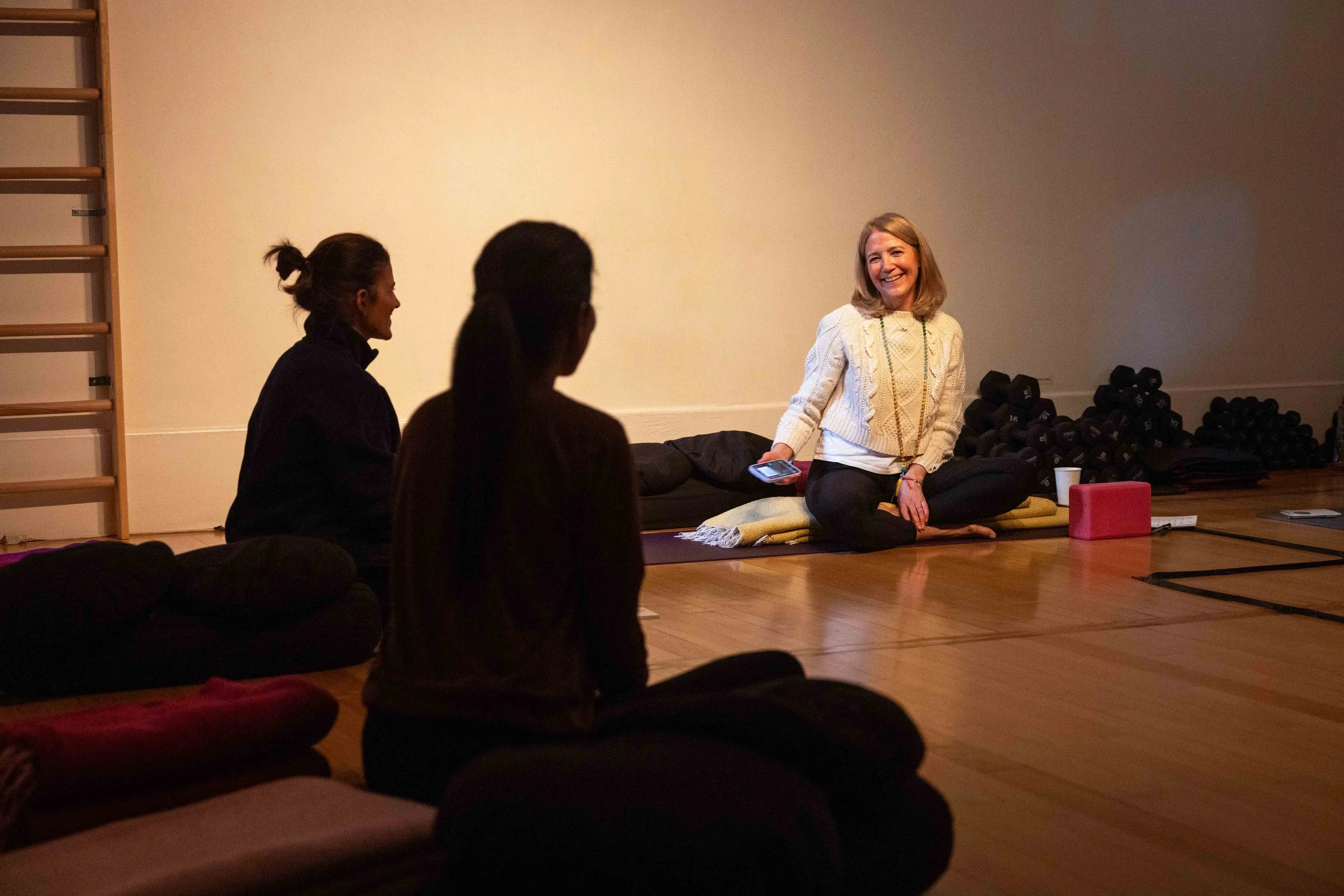 A woman smiling and sitting on a cushion in front of a group, holding a smartphone, in a room with a wooden floor, a wall with gym equipment, and a pile of dumbbells.