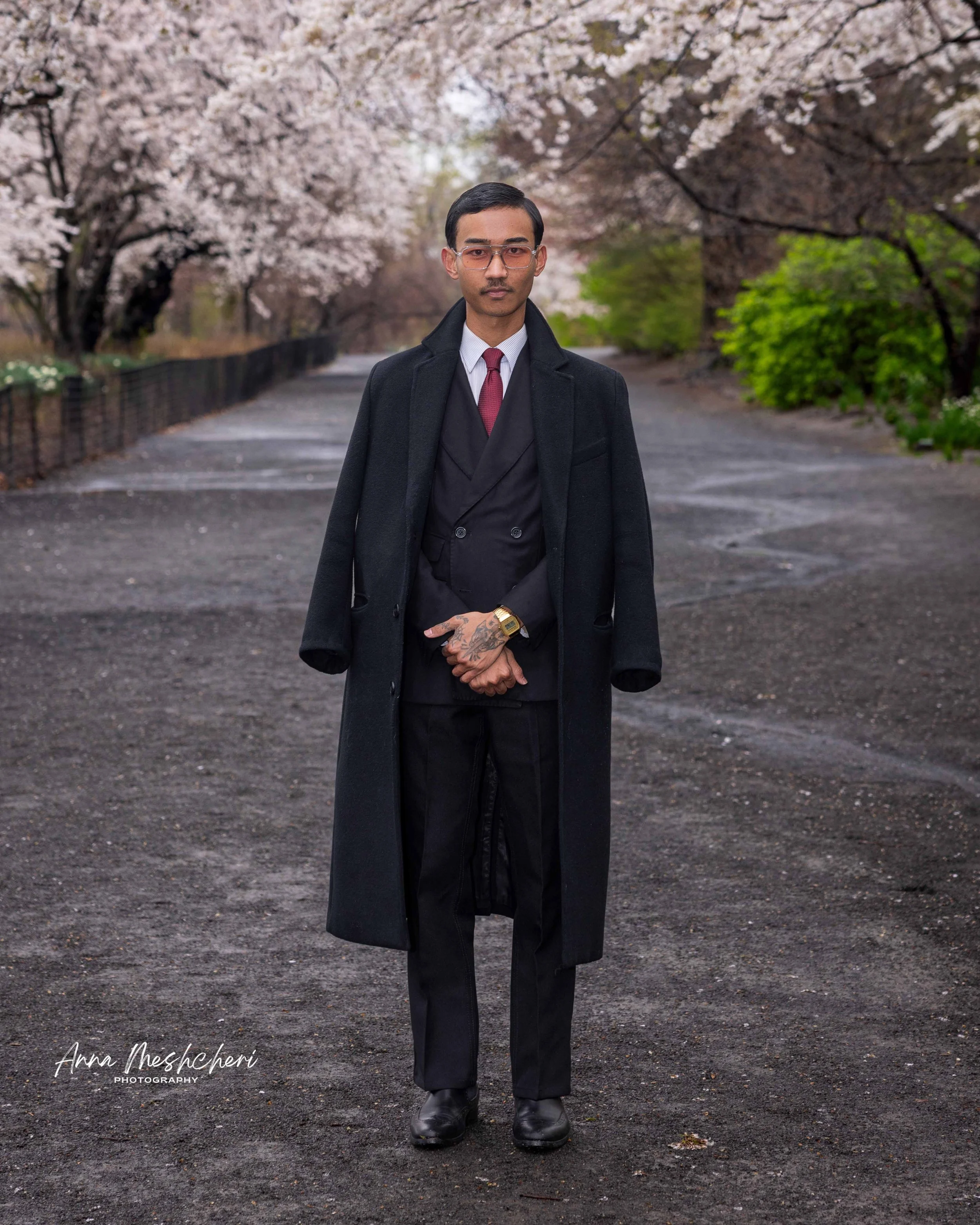 A man posing in a black suit and a black coat in Central Park, surrounded by cherry blossoms