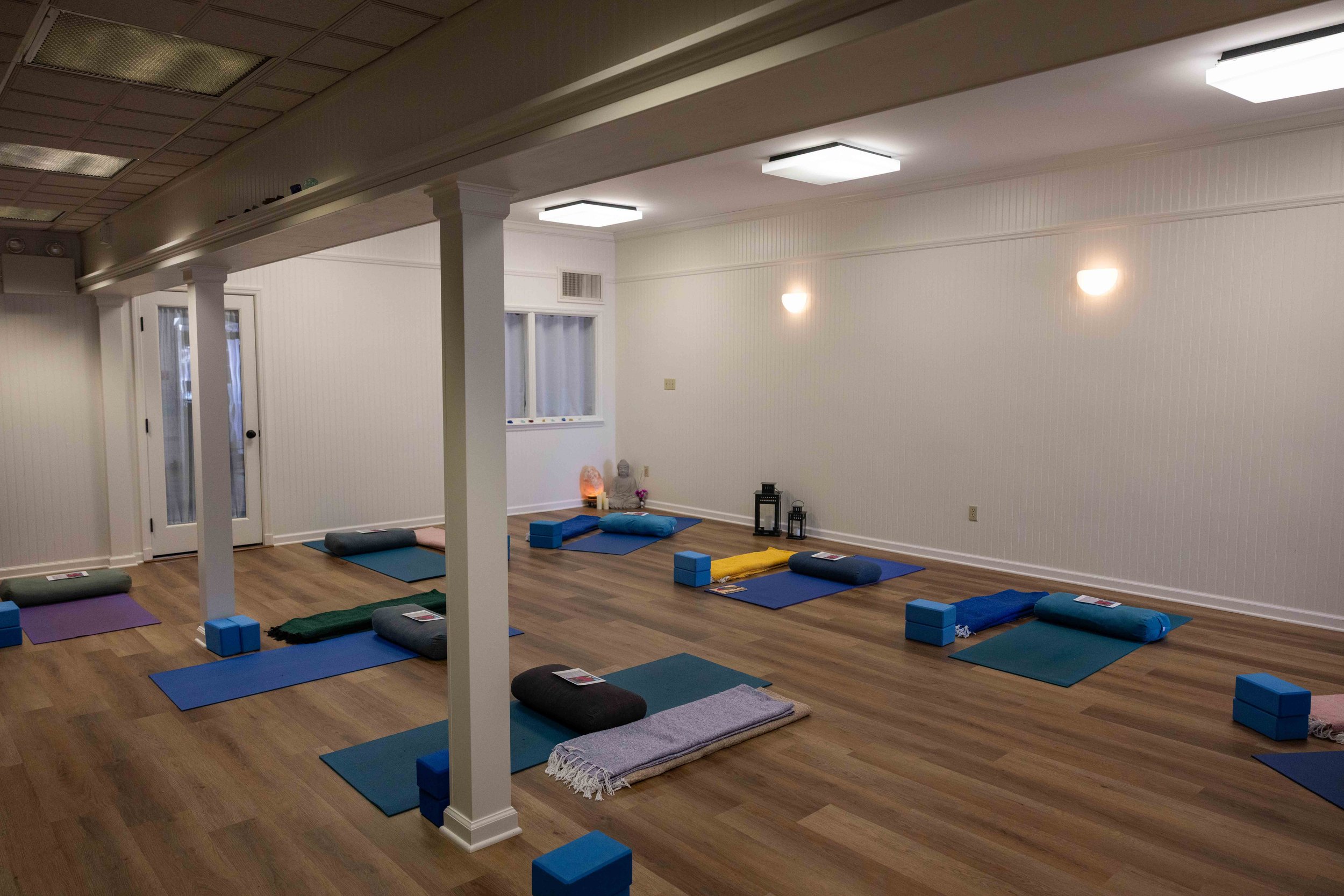 An indoor yoga or meditation studio with mats, bolsters, blocks, and blankets organized on a wooden floor. The room has white walls, a few windows, and ceiling lights, with a small Buddha statue and decorative lanterns in the background.