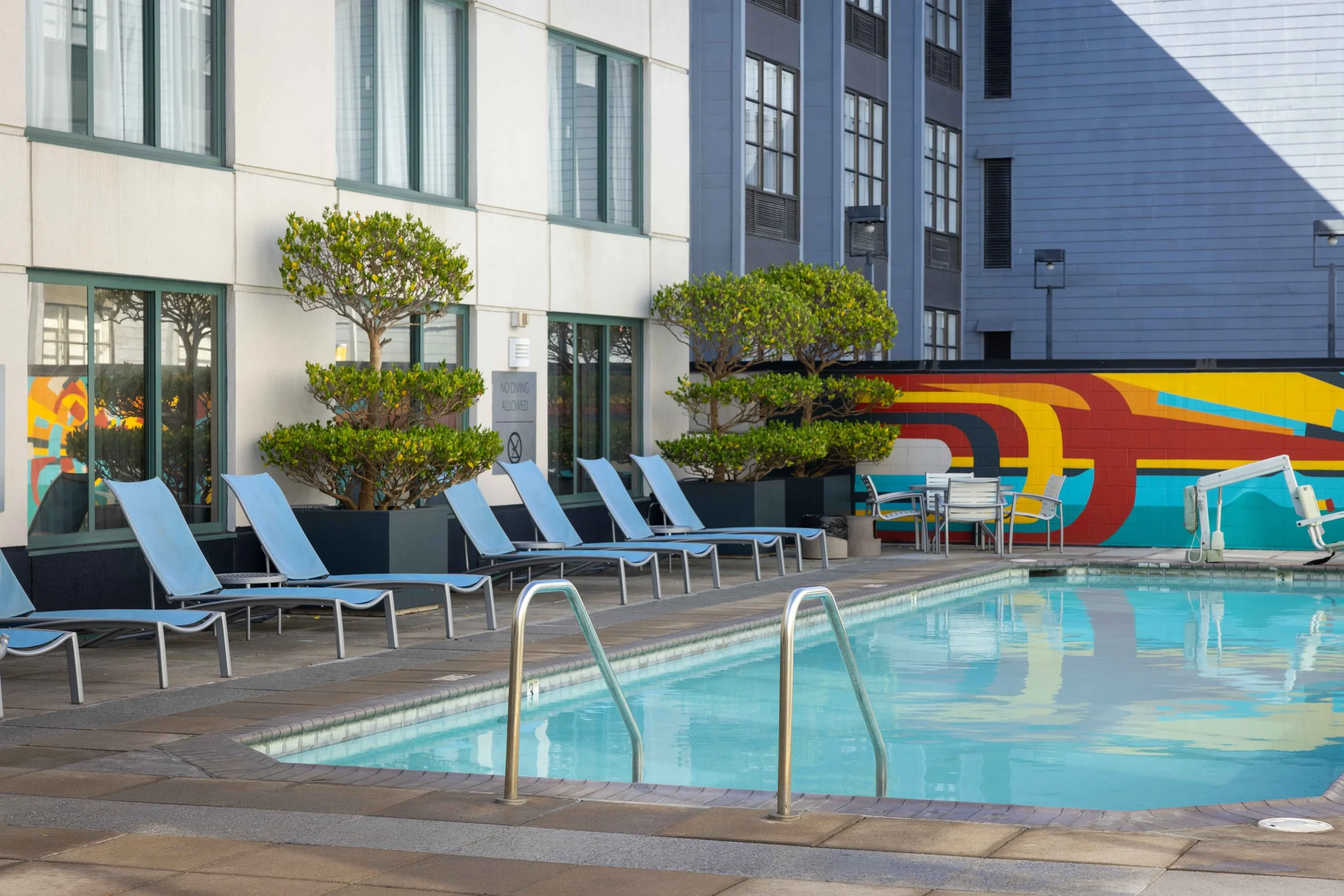 Empty swimming pool with lounge chairs and colorful mural on wall, modern buildings in background, trees by building, sign indicating no diving allowed.