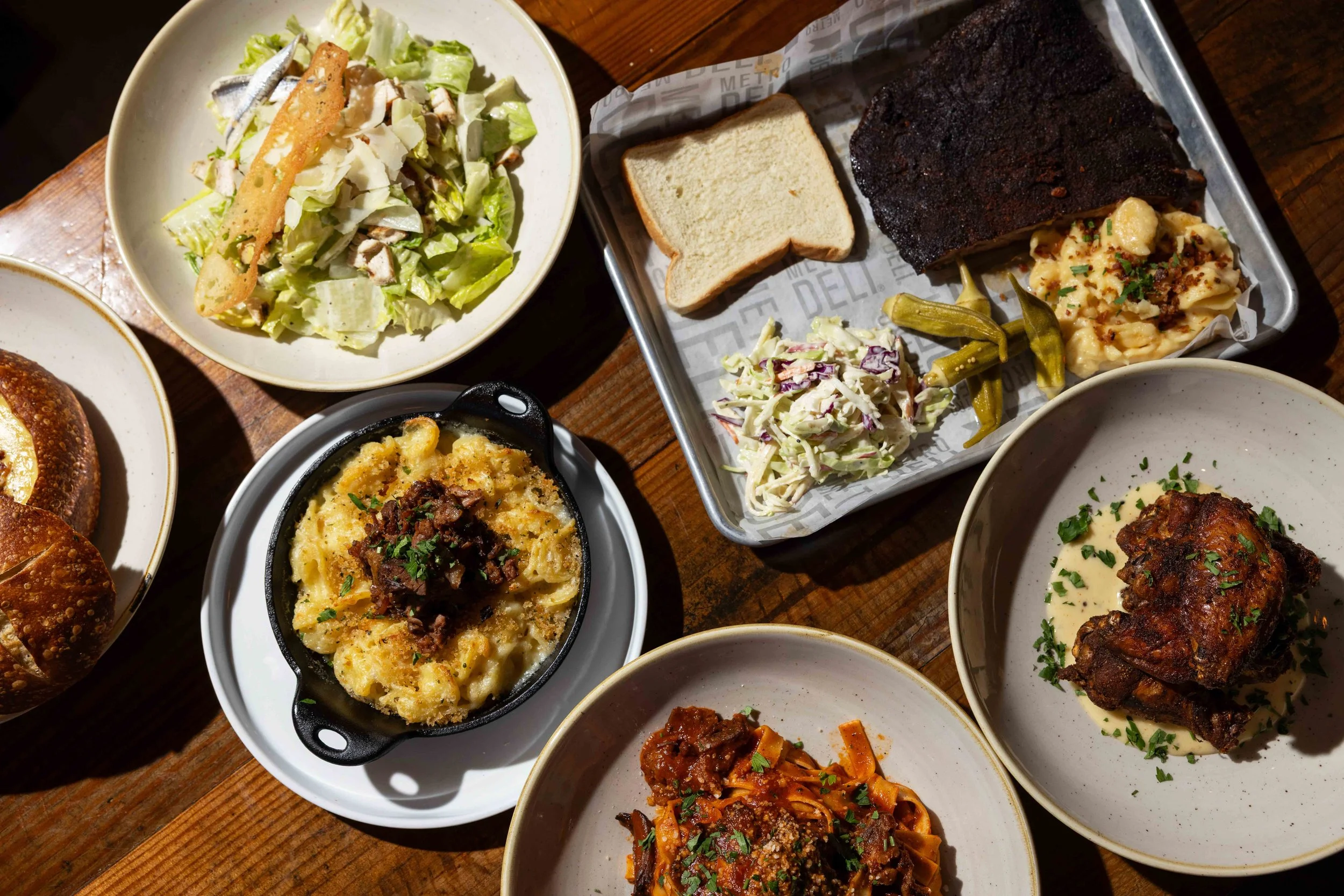 Assorted dishes of macaroni and cheese, coleslaw, baked casserole, sliced bread, ribs, and salad on a wooden table.