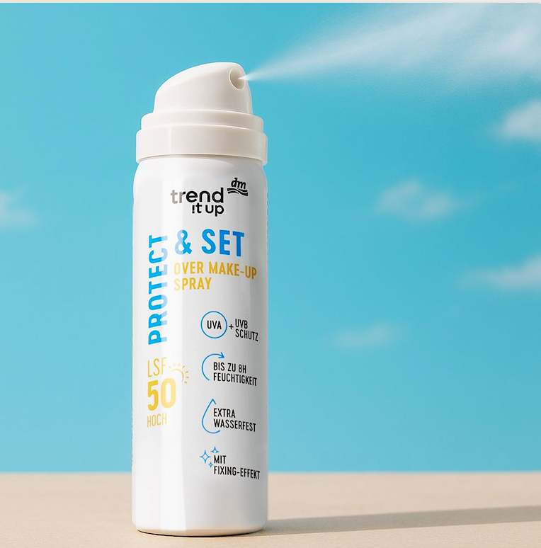A white spray can with blue and orange text labeled 'Protect & Set Over Makeup Spray' on a sandy surface with a blue sky background, spraying a mist.
