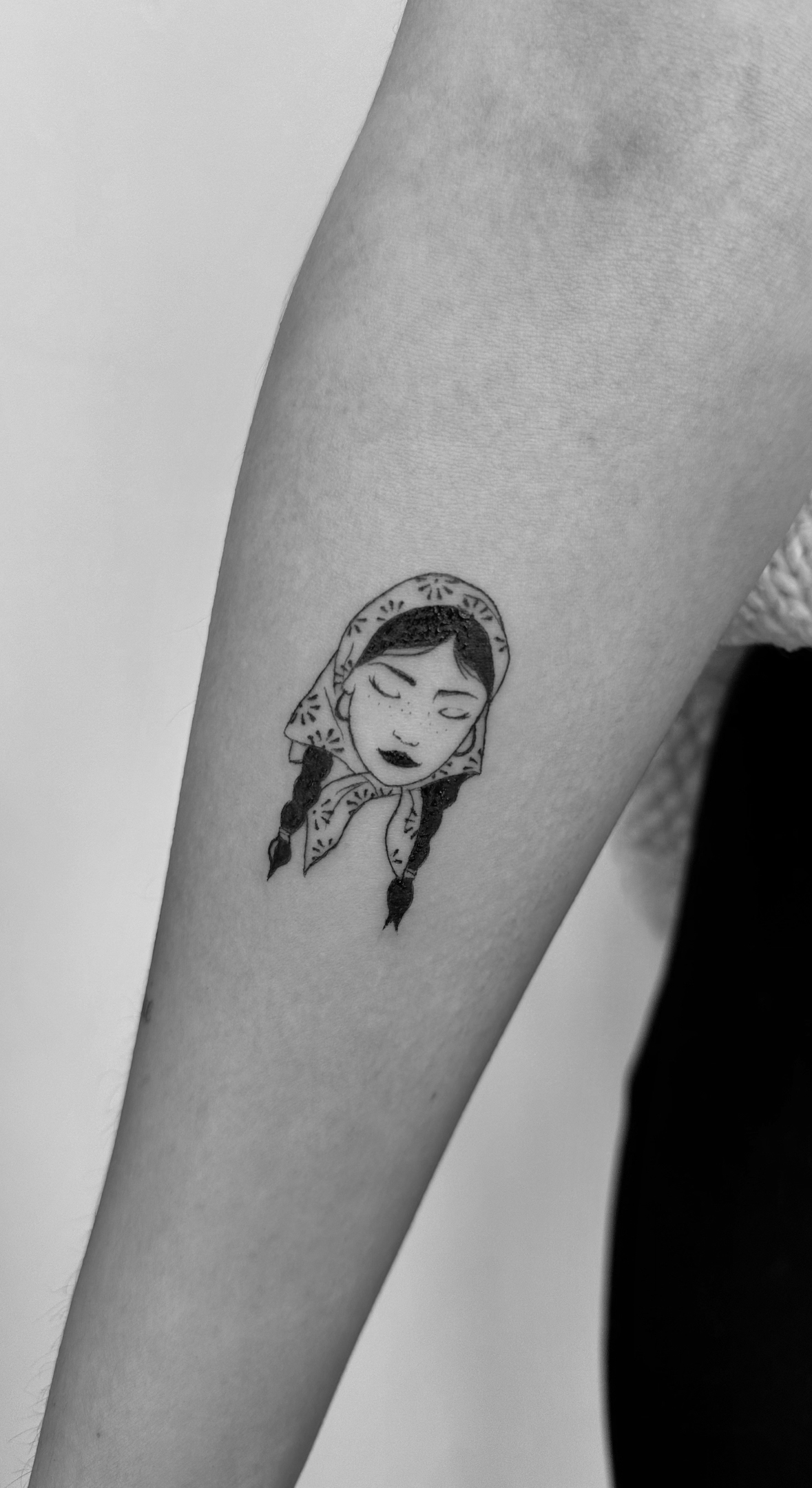 Black and white tattoo of a woman with braided hair and a headscarf on a person's arm.