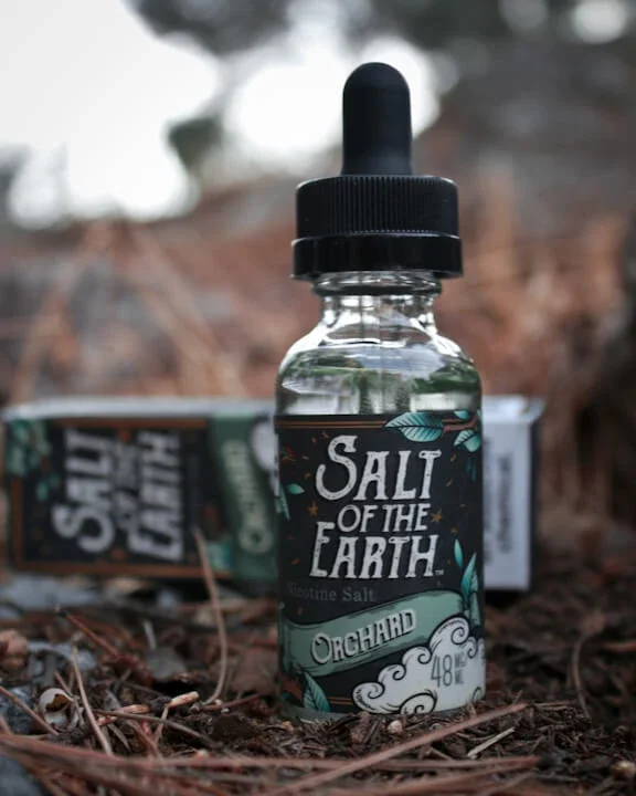 A glass dropper bottle labeled 'Salt of the Earth' with 'Gourmet Salt' and 'Orchard' written on it, placed outdoors on the ground surrounded by pine needles and twigs, with a blurred container in the background.