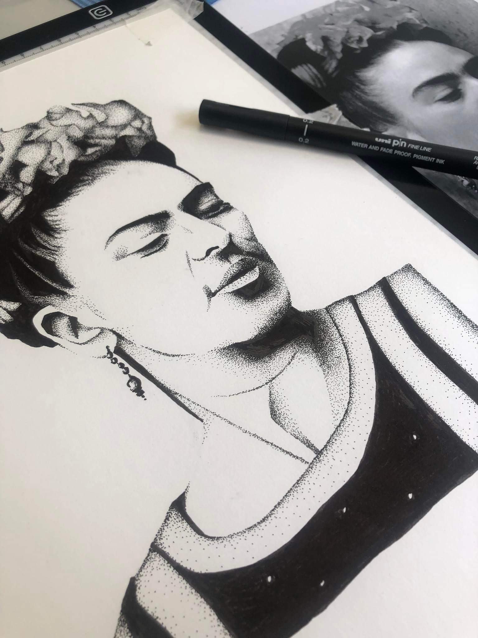 A black and white stippled portrait of a woman with her eyes closed, wearing earrings, and a striped top, with art supplies and a reference photo nearby.