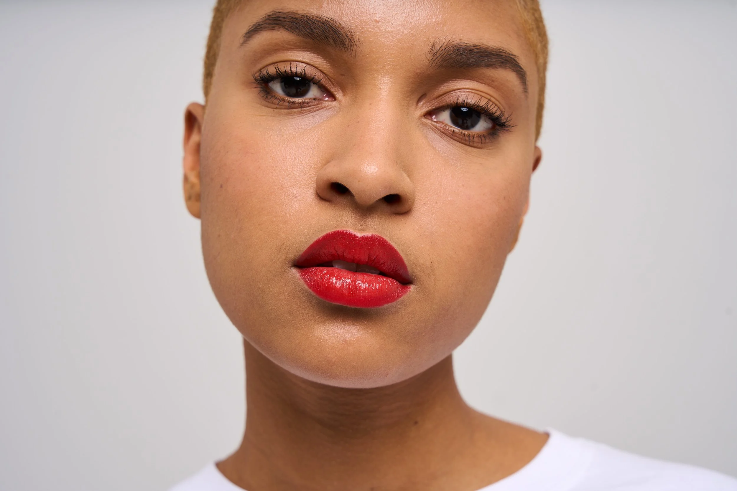Close-up of a woman with light brown skin, short blonde hair, red lipstick, and light makeup, looking directly at the camera against a plain light background.