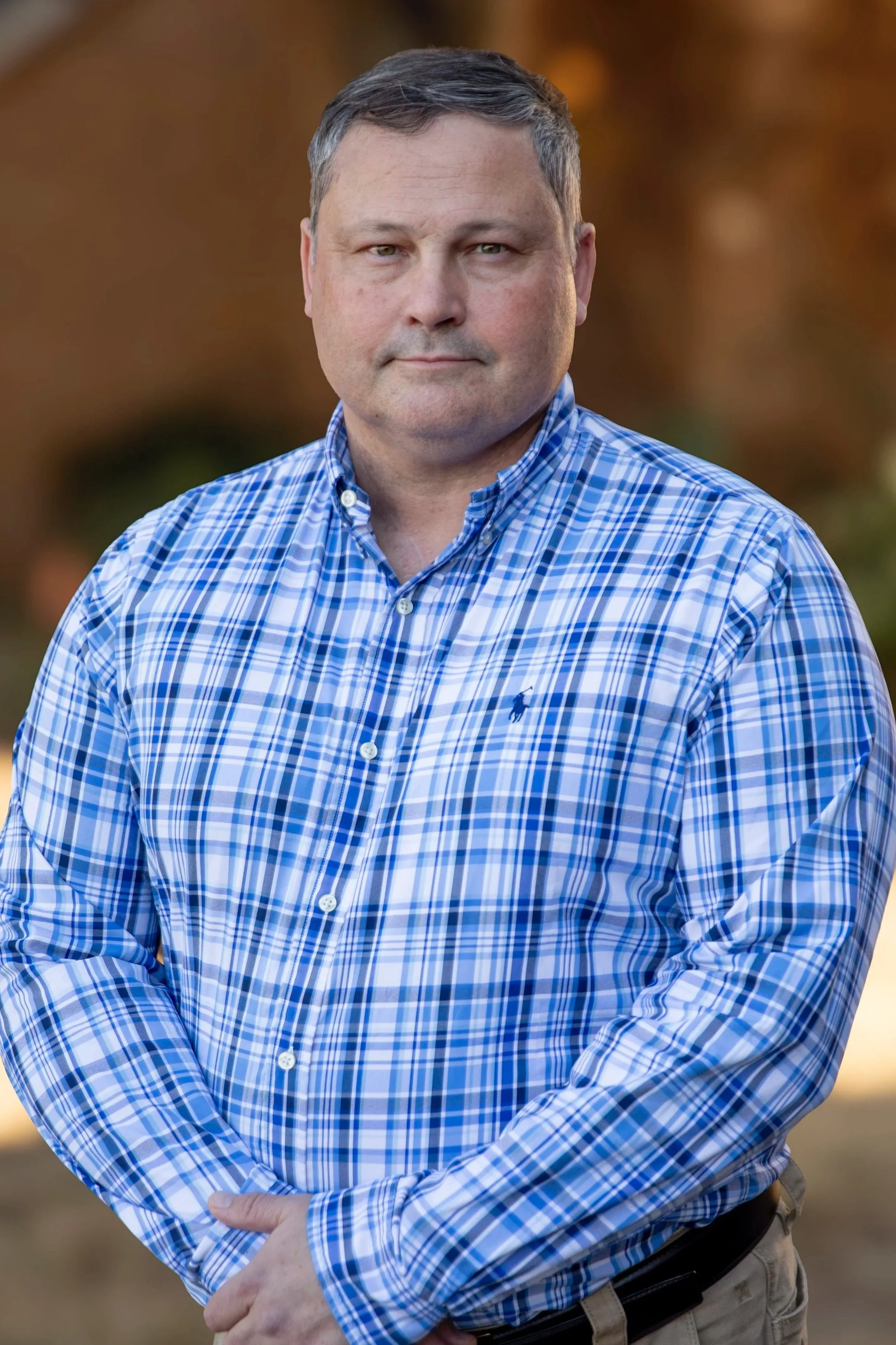 A portrait of a middle-aged man with short brown hair, wearing a dark suit jacket over a light blue dress shirt, smiling with a neutral gray background.