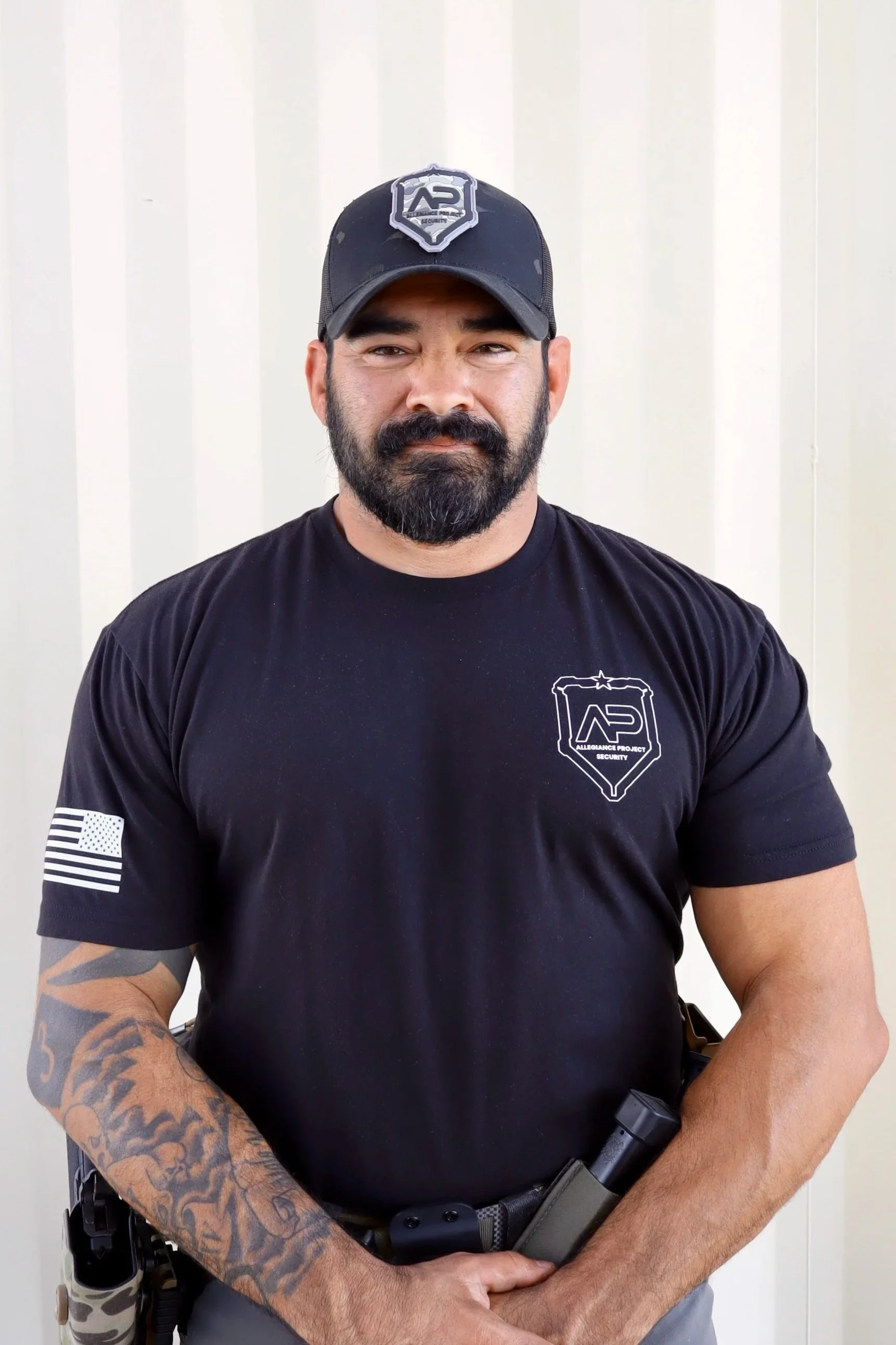 A man with a beard and sunglasses standing outdoors in front of a shooting range setup with paper targets, trees in the background, wearing a black t-shirt and khaki pants.