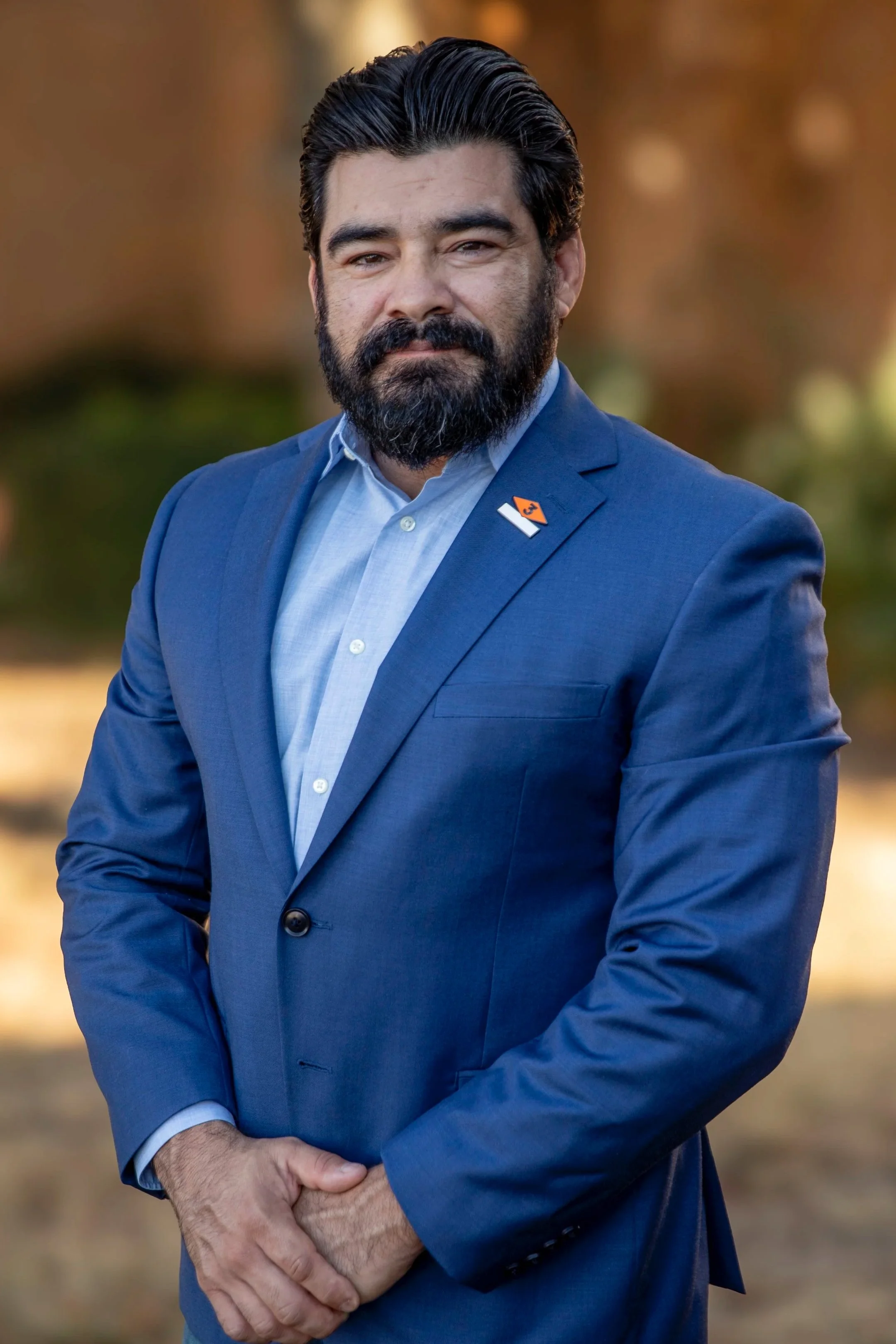A man with a beard and sunglasses standing outdoors in front of a shooting range setup with paper targets, trees in the background, wearing a black t-shirt and khaki pants.
