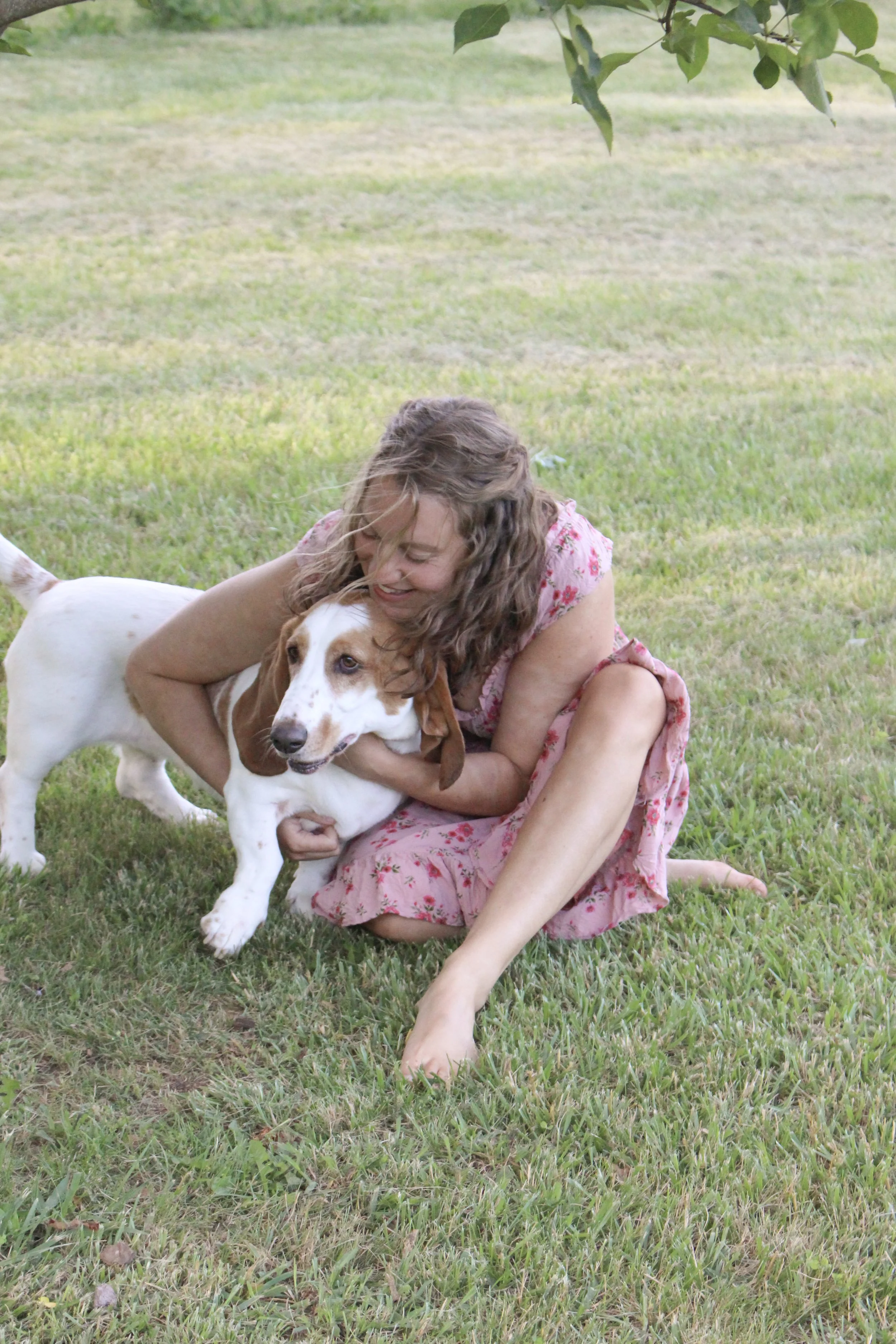 A woman in a pink floral dress hugging a Basset Hound dog on a grassy lawn.
