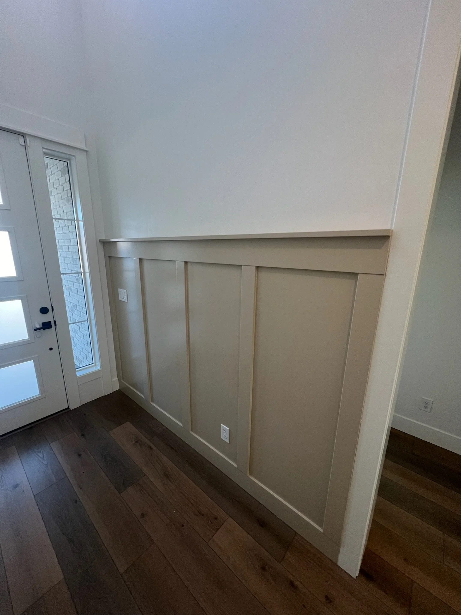Interior view of a corner with a white wall, a white door with glass panels, a white wall with wood paneling, and dark hardwood flooring.