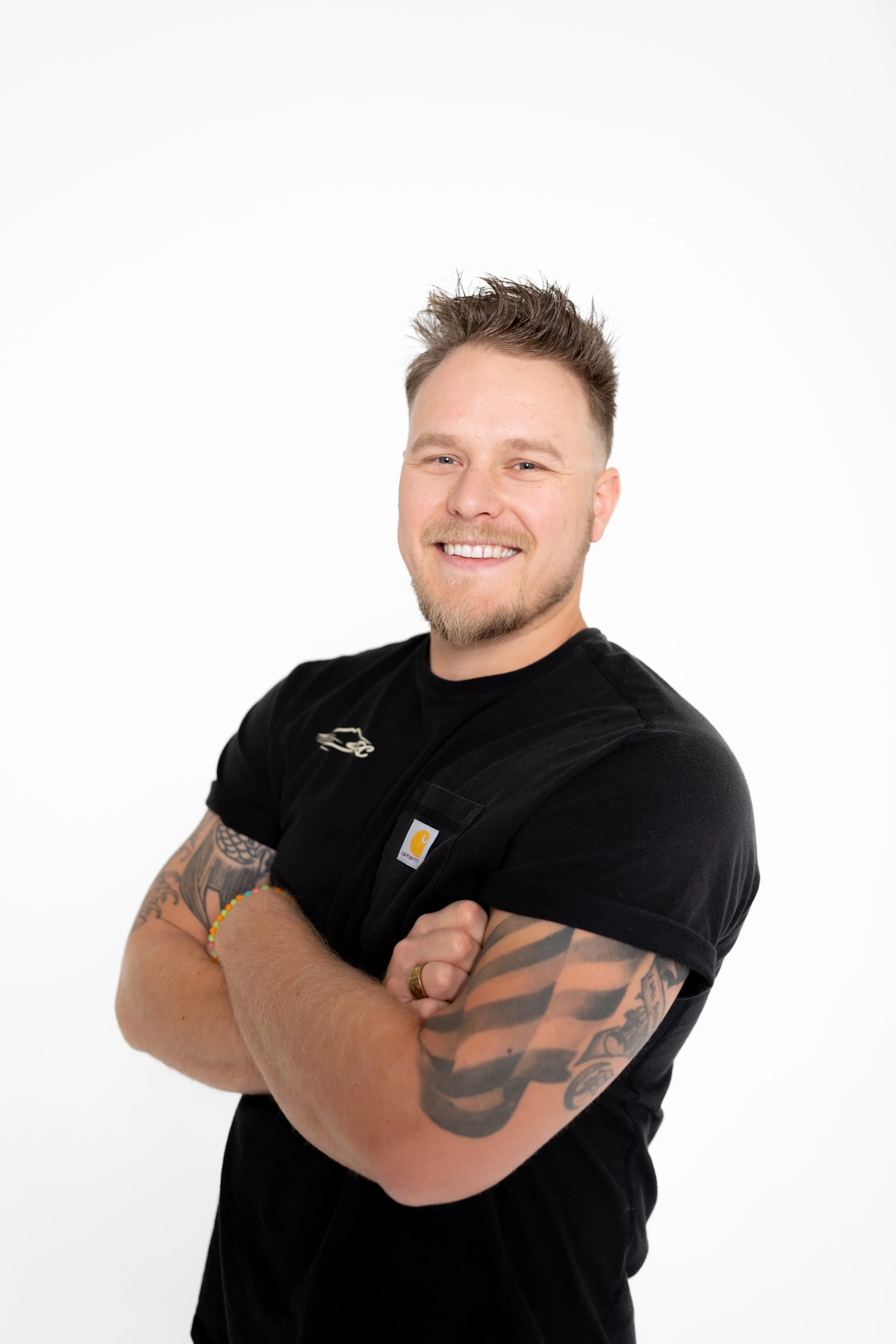 A young man with short, styled hair and a beard, smiling with arms crossed, wearing a black T-shirt with visible tattoos on his arms against a plain white background.