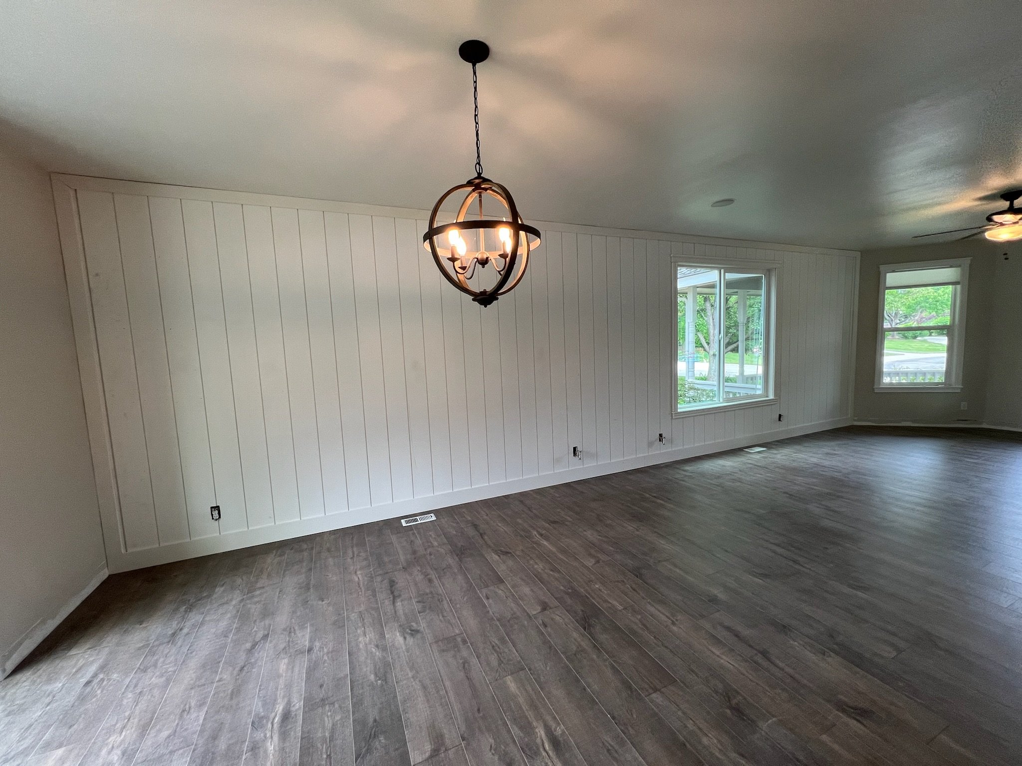 Empty living room with white paneled walls, dark wood flooring, large windows, and a modern chandelier hanging from the ceiling.