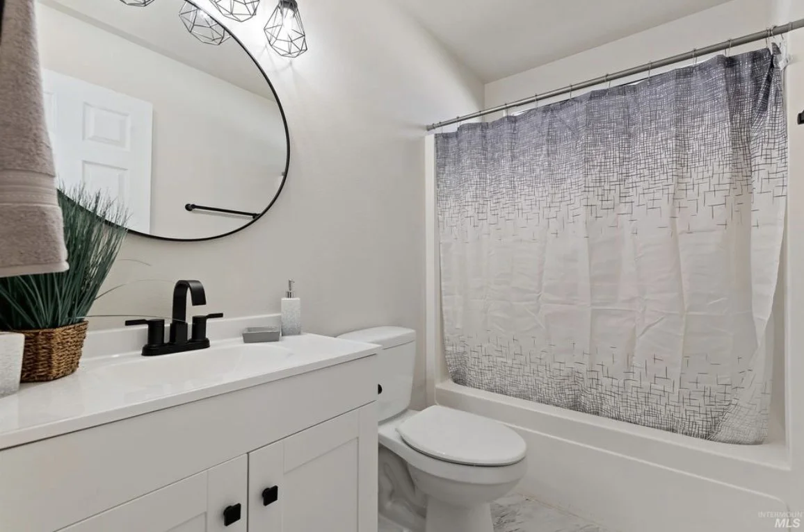 Bathroom with white vanity, round mirror, black fixtures, decorative plant, soap dispenser, and a bathtub with a geometric patterned shower curtain.