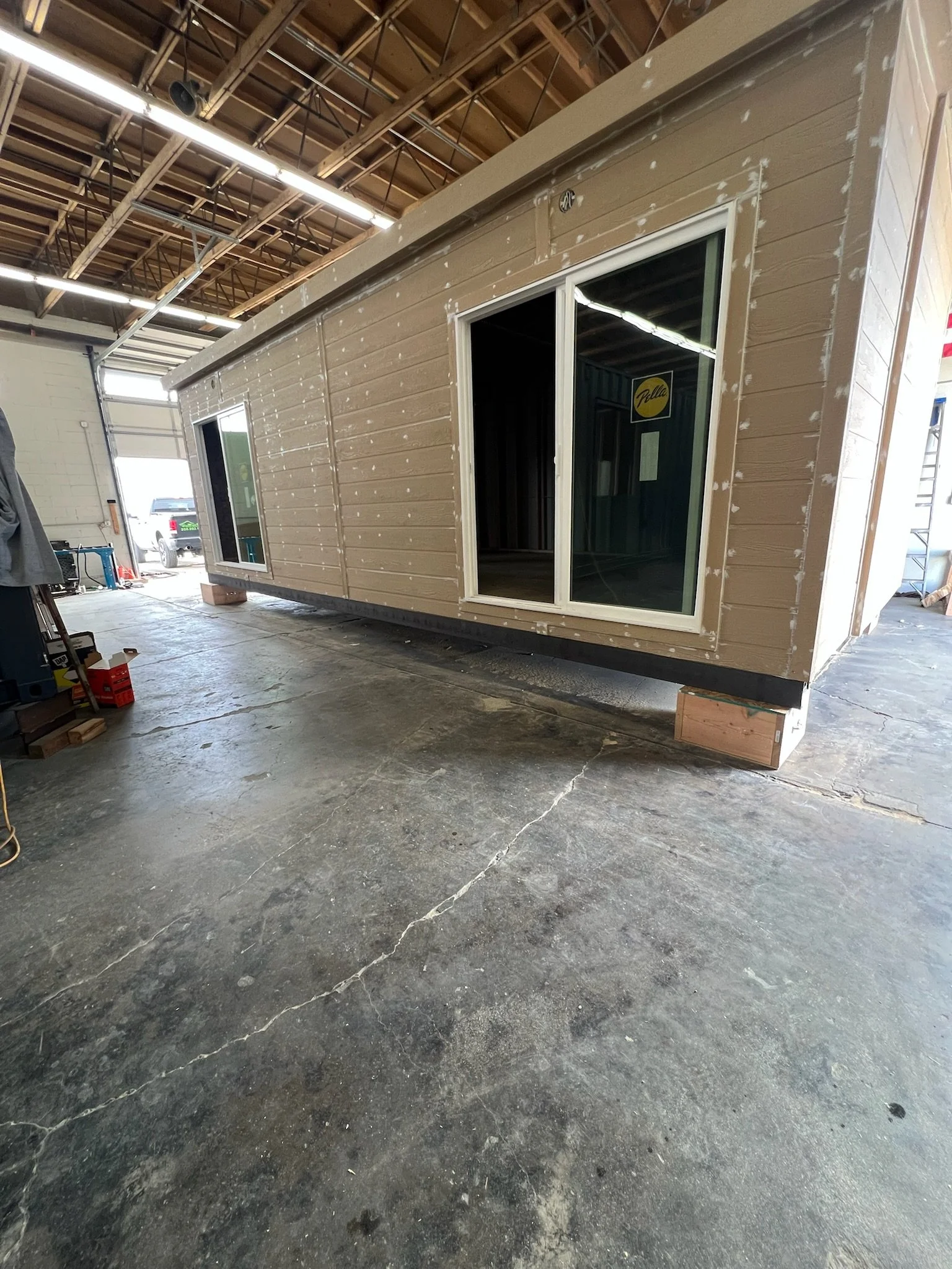 Interior of a warehouse with a small wooden structure that has a large window and siding, set on a concrete floor with some crates and tools around.