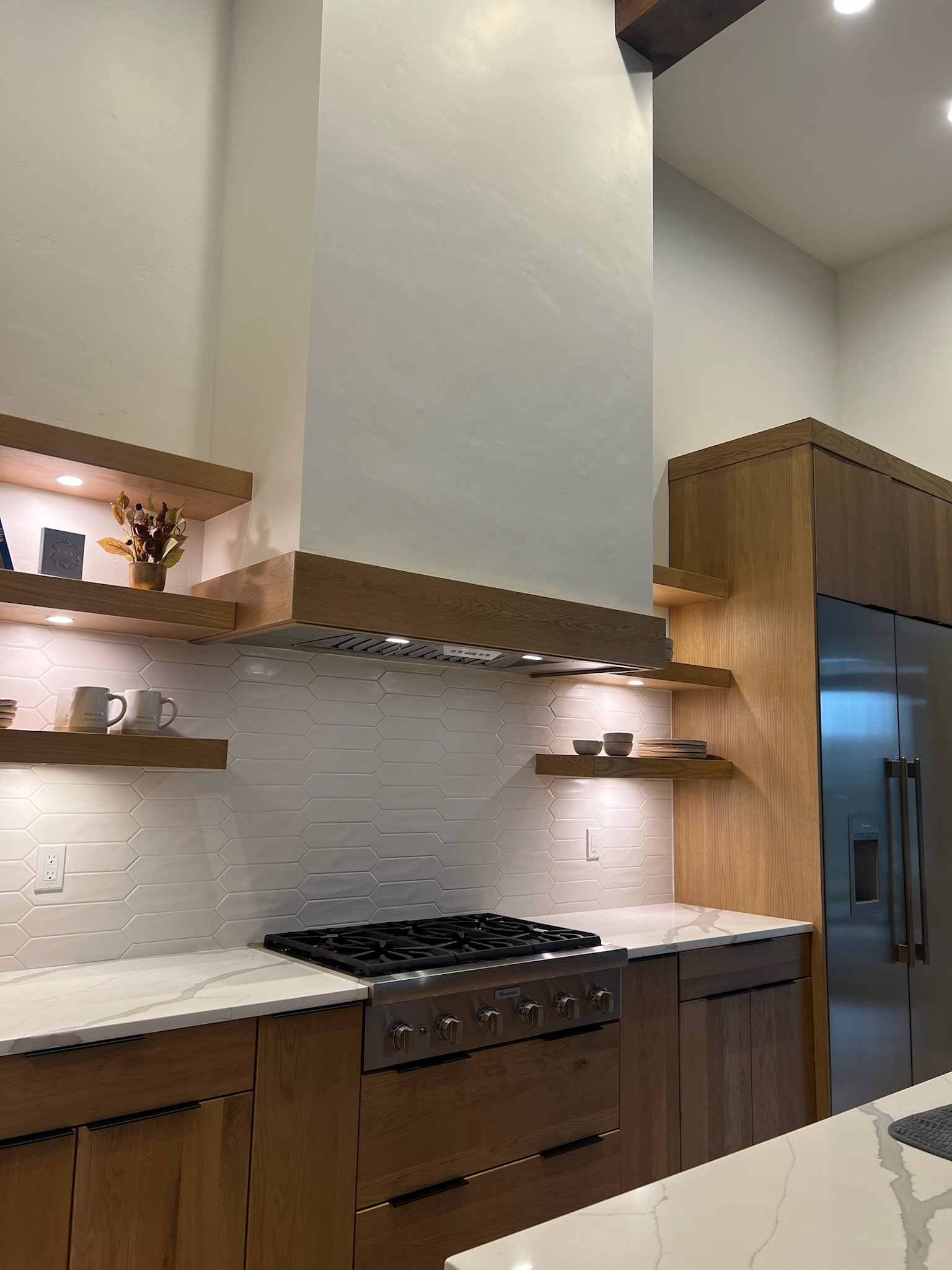 Modern kitchen with white hexagonal tile backsplash, wooden cabinets, stovetop, open floating shelves with decorative items, and stainless steel refrigerator.