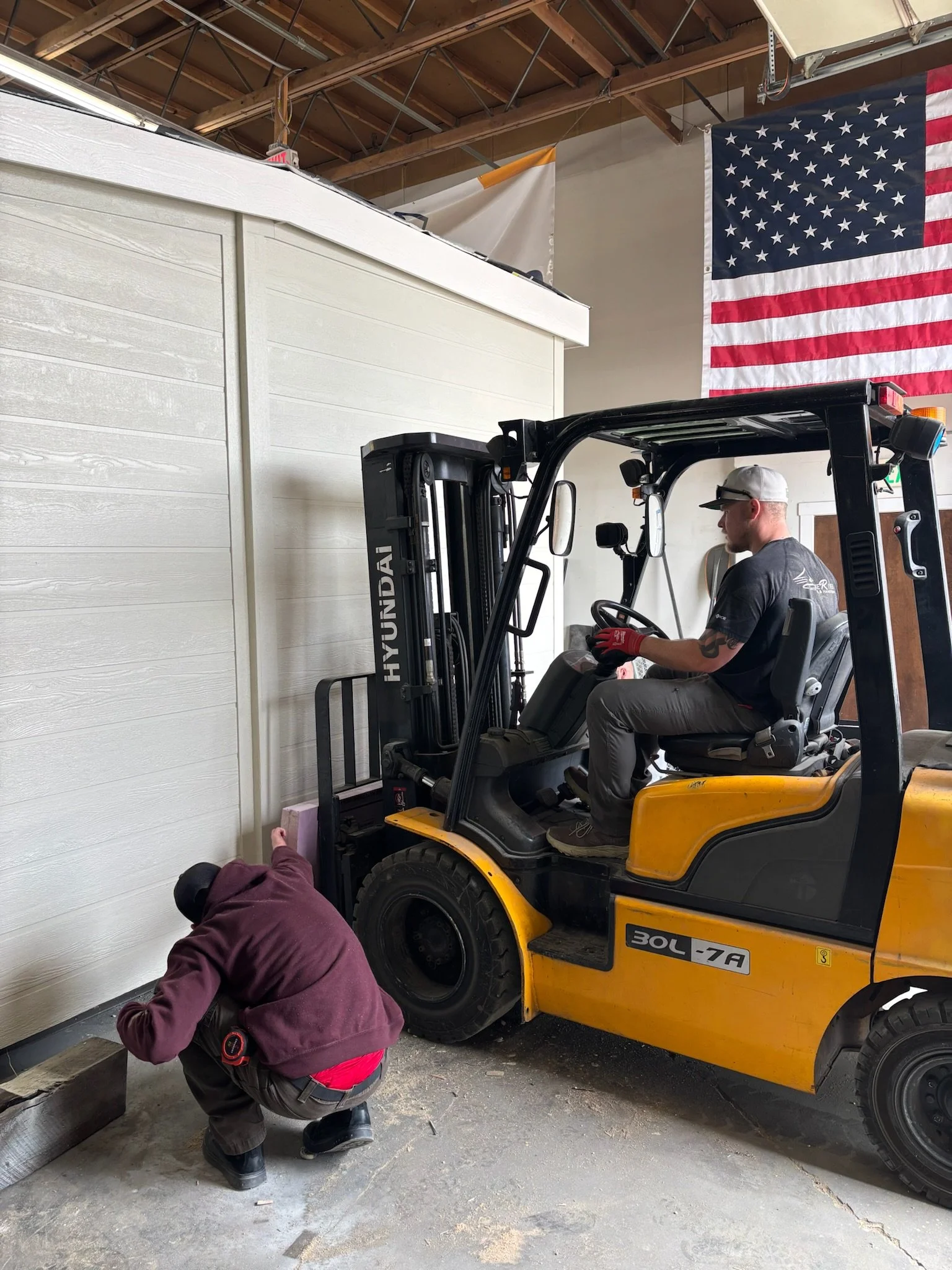 Two people working with a forklift inside a building with an American flag on the wall and a white panel wall. One person is operating the forklift, and the other is kneeling on the floor near a block of wood.