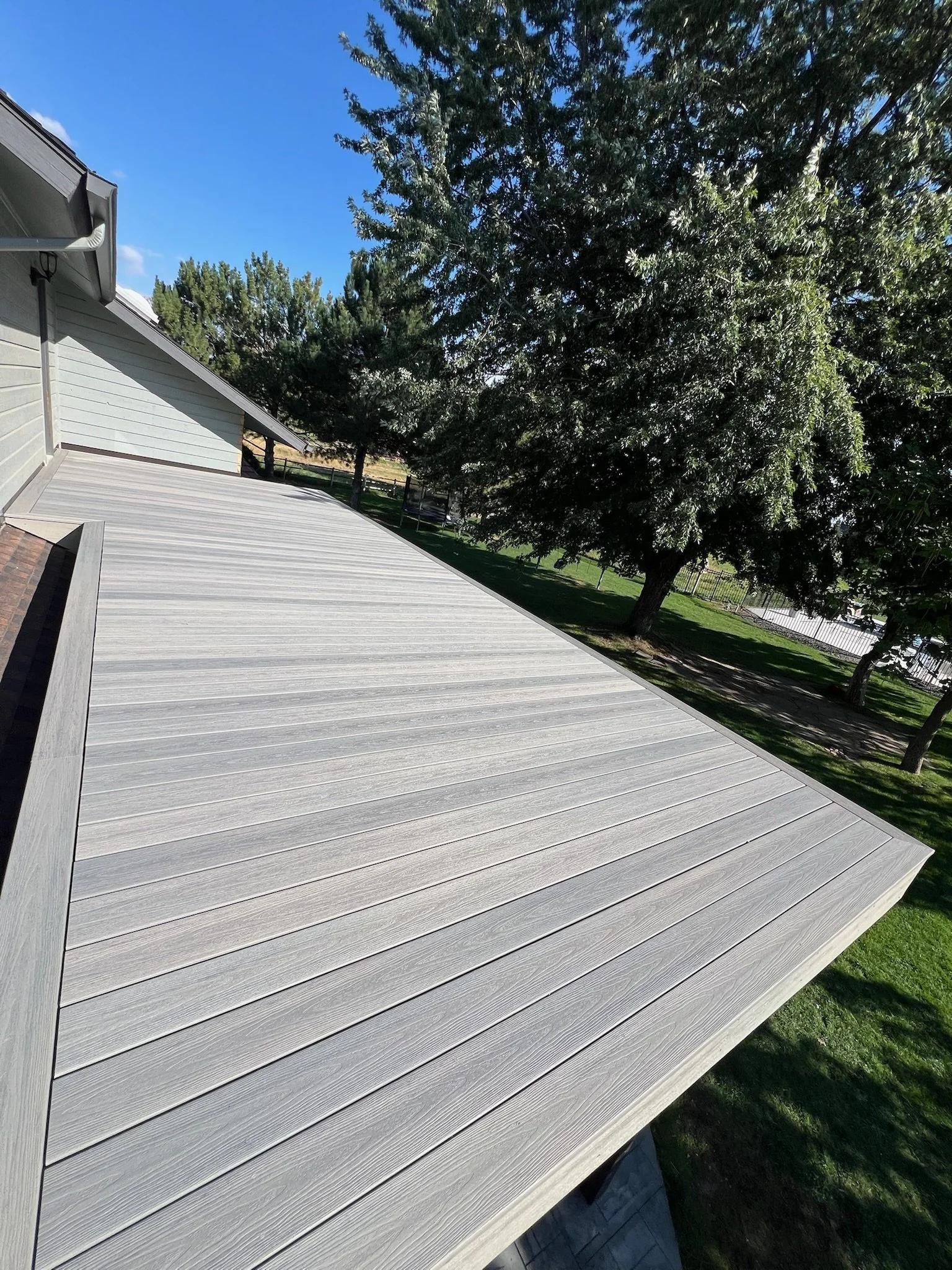A modern, flat, gray wooden deck attached to a house with light gray siding, surrounded by green grass and tall trees under a clear blue sky.