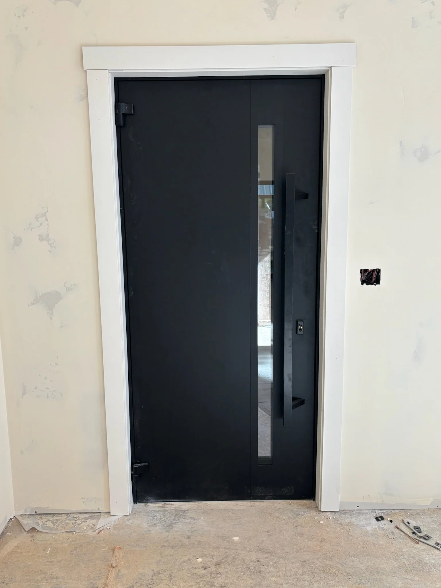 Black door with a long vertical window and black handle, surrounded by white trim, in a room with unfinished drywall and construction debris on the floor.