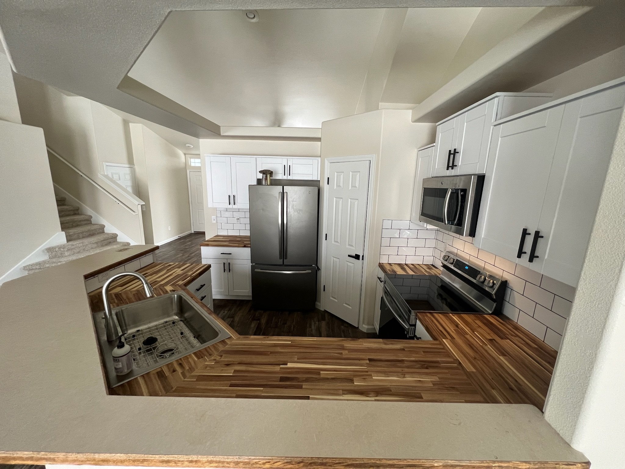 Interior view of a modern kitchen with white cabinets, stainless steel appliances, a wooden countertop, and a staircase nearby.