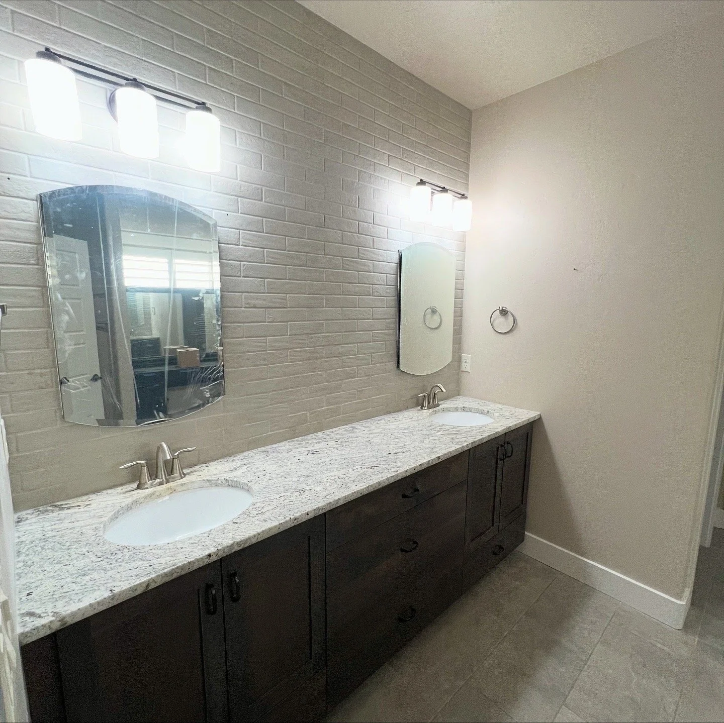 Bathroom with dual sinks, granite countertop, dark wood cabinets, brick accent wall, two mirrors, and modern light fixtures.