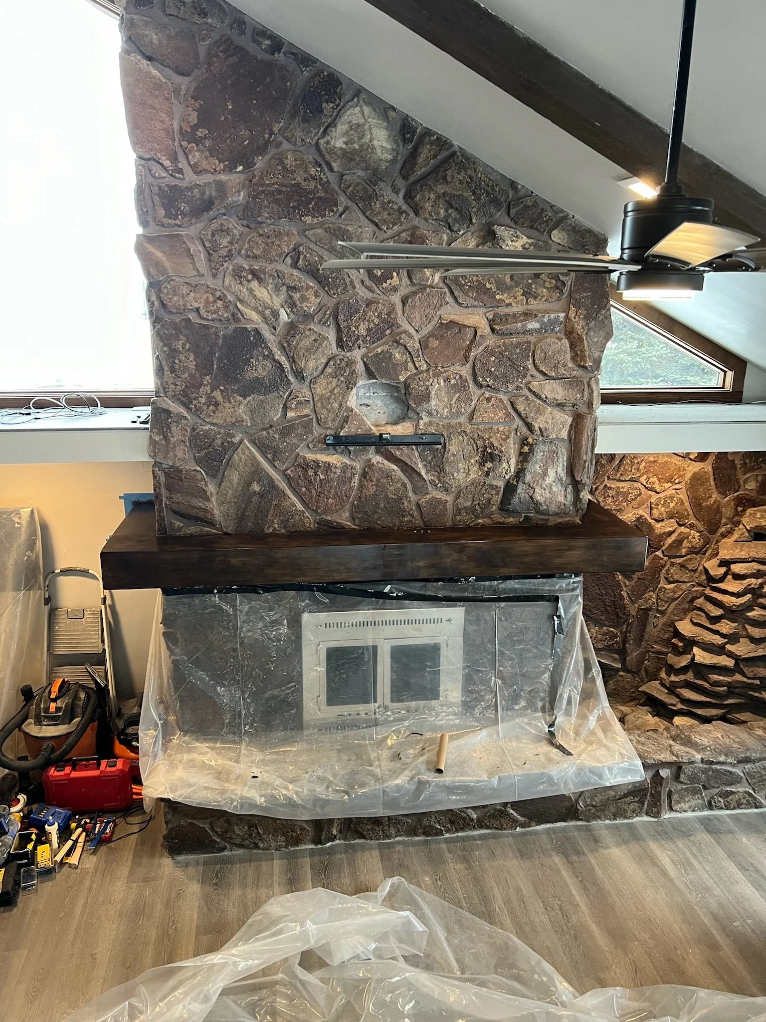 A stone fireplace with a black two-sided fire grate in front of it, partially covered with plastic sheeting. To the left, there is a window with a window ledge, and on the floor, various tools and equipment are arranged, including a red toolbox, a va