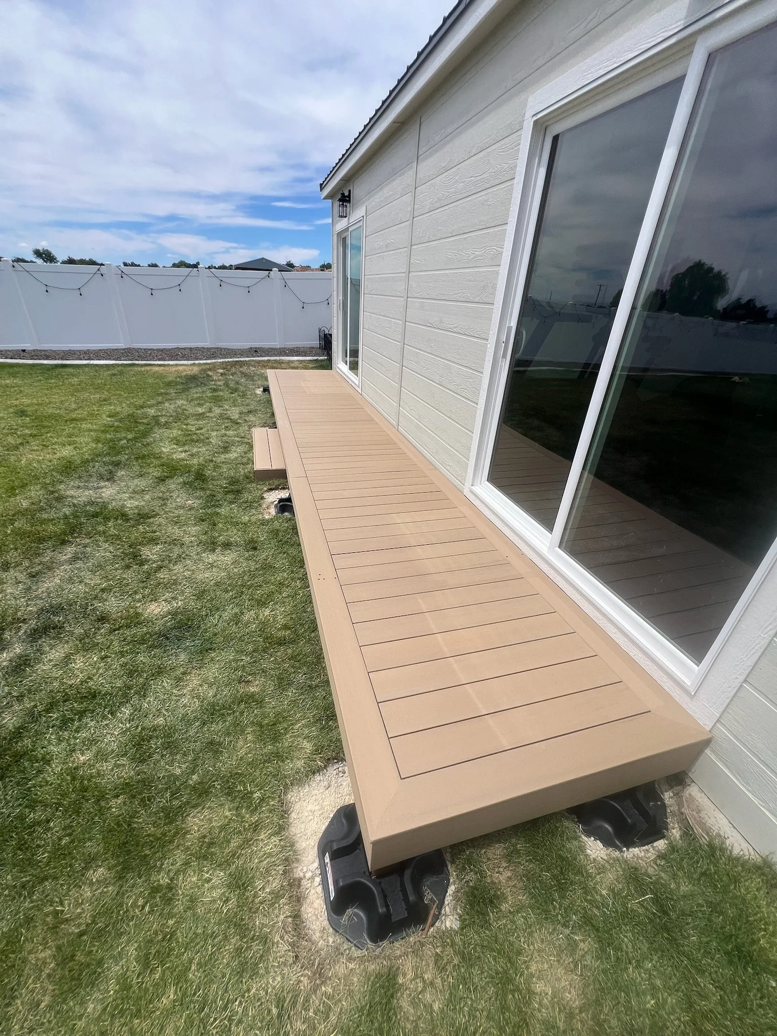 Newly installed wooden deck attached to the side of a house with beige siding. The deck is above ground and supported by black stands. There are sliding glass doors leading to the house, and a black outdoor wall-mounted lantern light fixture nearby. 