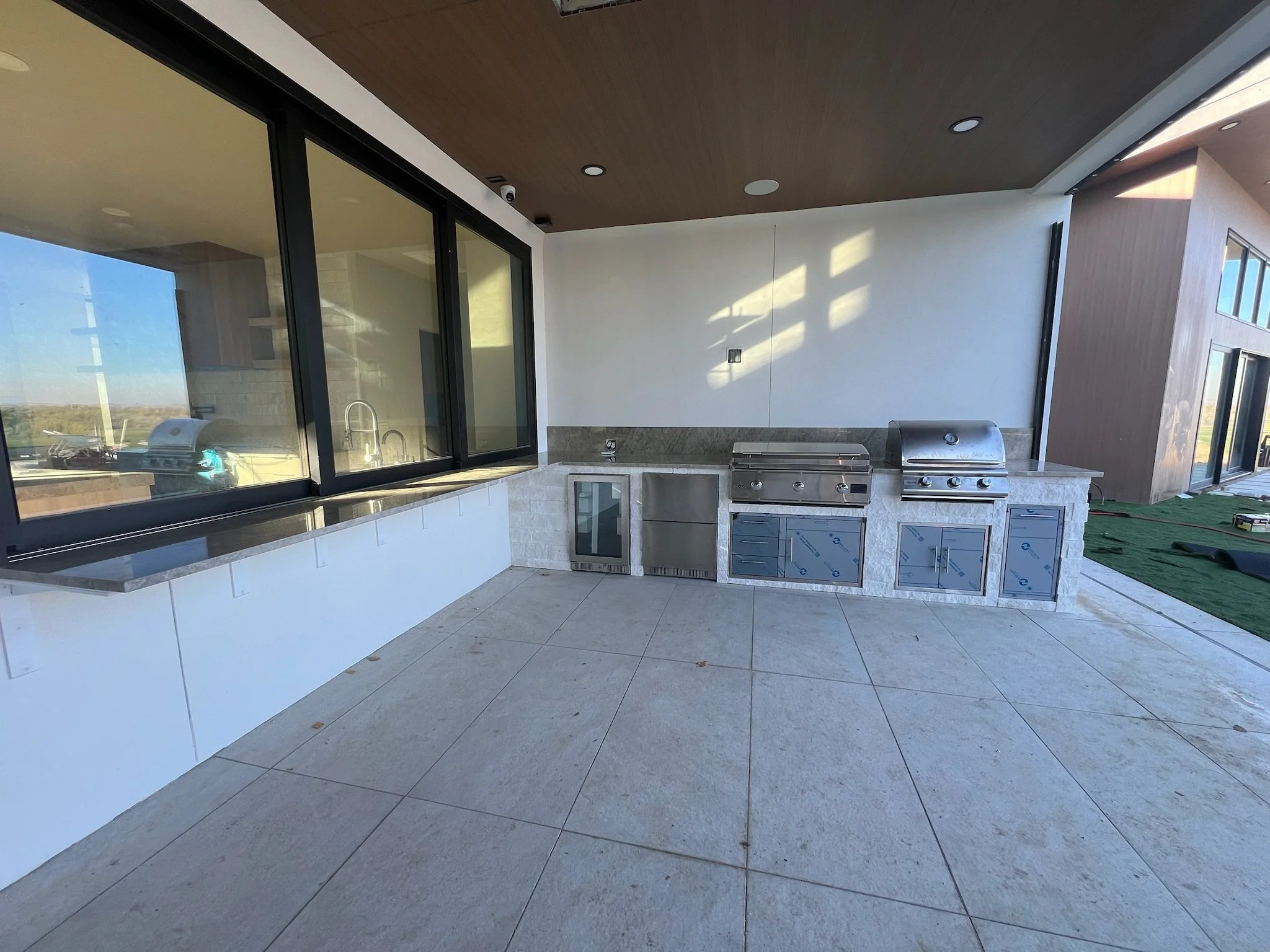 Modern outdoor kitchen with stainless steel grill, white cabinets, and large windows with view of outdoor landscape.
